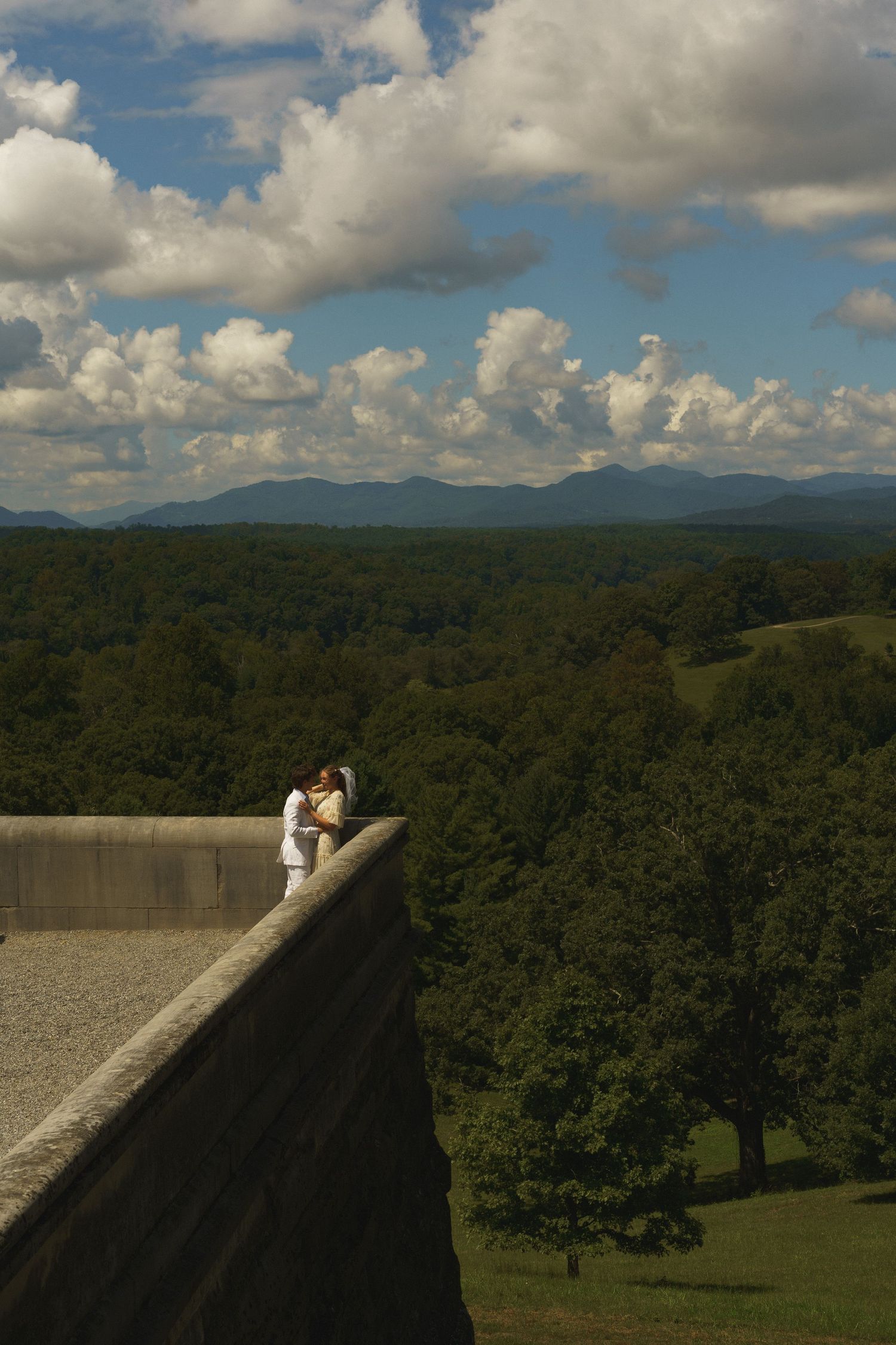 A couple stands on a scenic overlook with mountains and forests stretching to the horizon.