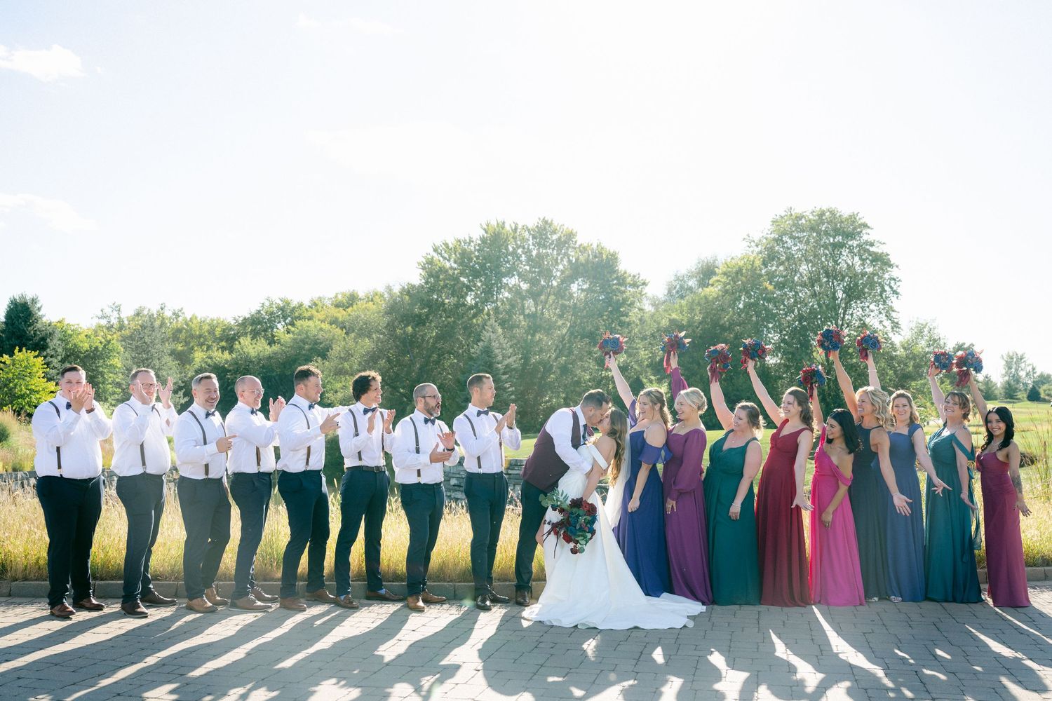Bright sunlit outdoor wedding party photo with bridesmaids in colorful dresses and groomsmen in light shirts lined up.