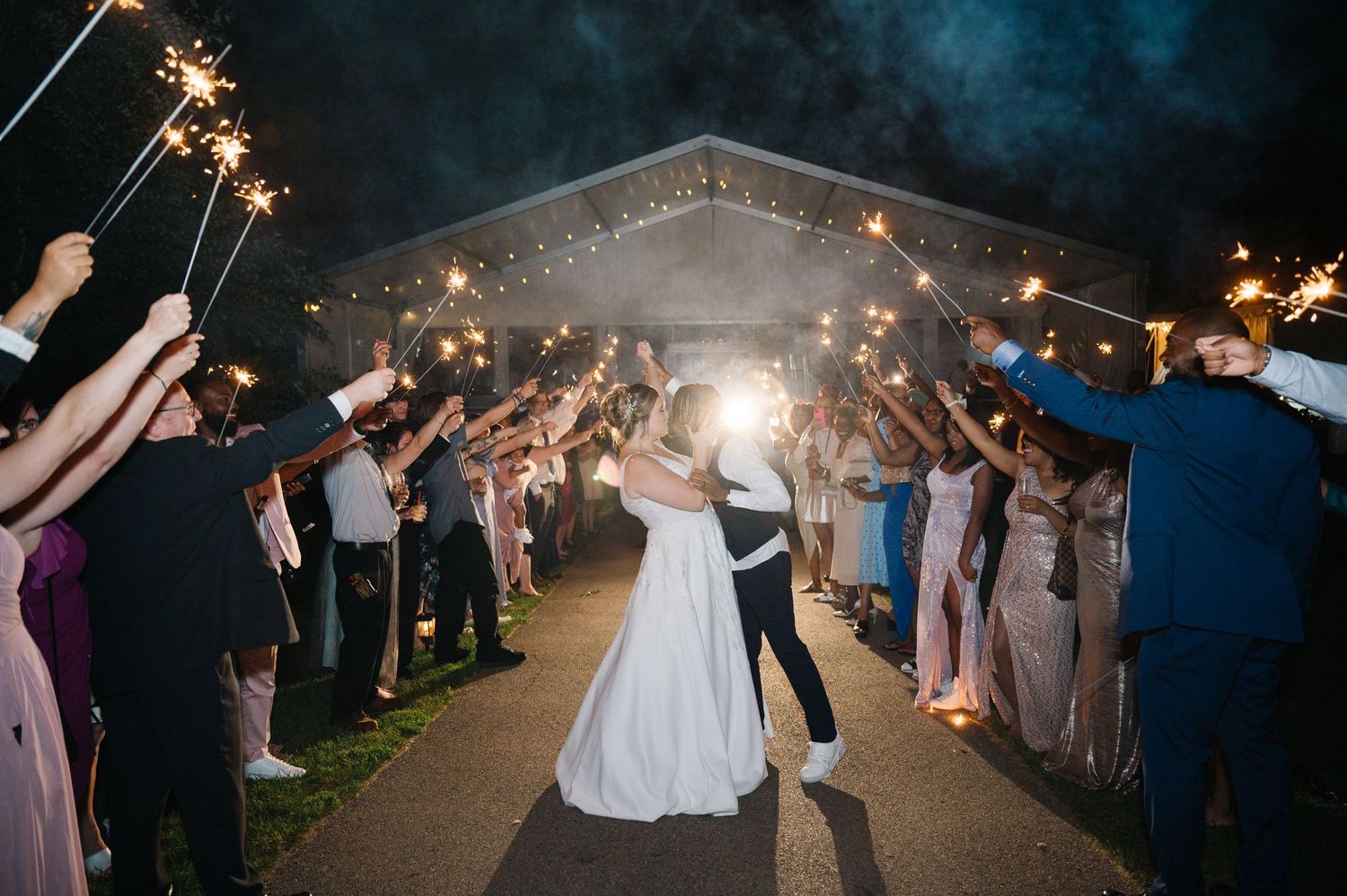 Wedding guests create a sparkler tunnel exit for newlyweds dancing under the night sky at outdoor reception.