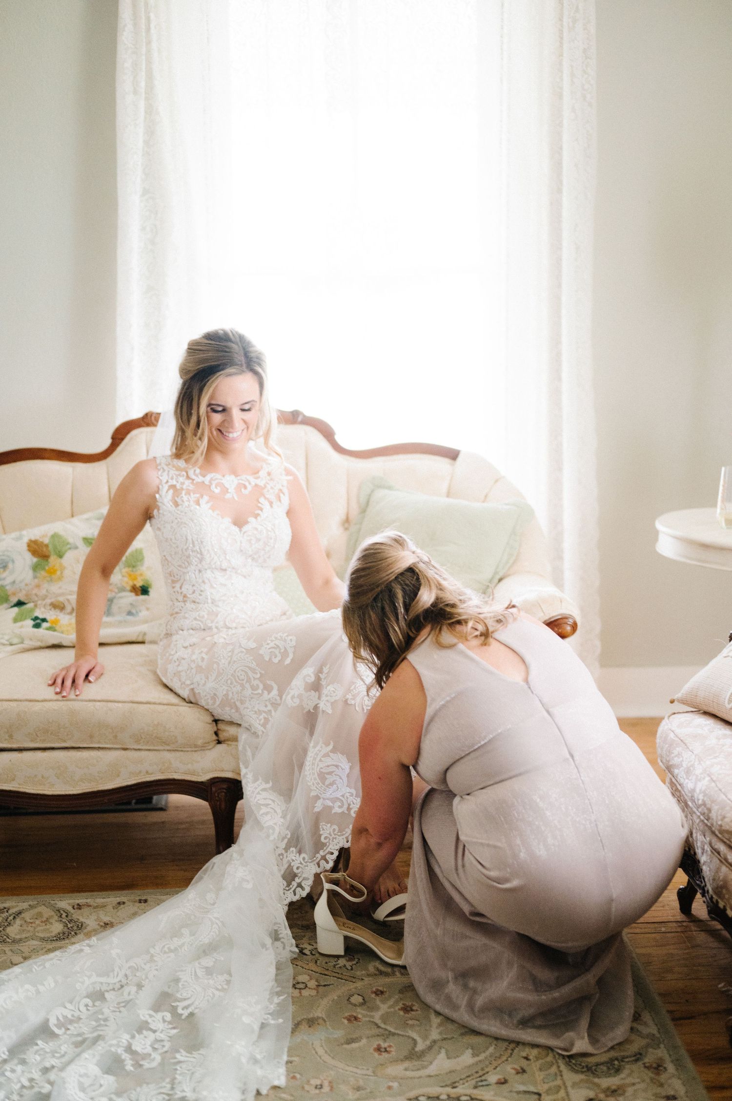 Wedding dress preparation scene with delicate lace details in elegant light-filled room with antique furniture.