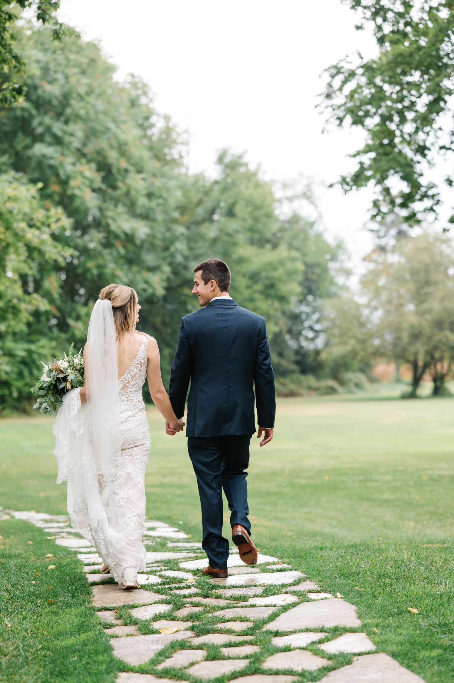Newlywed couple walking along a stone garden path surrounded by lush green trees and grass.