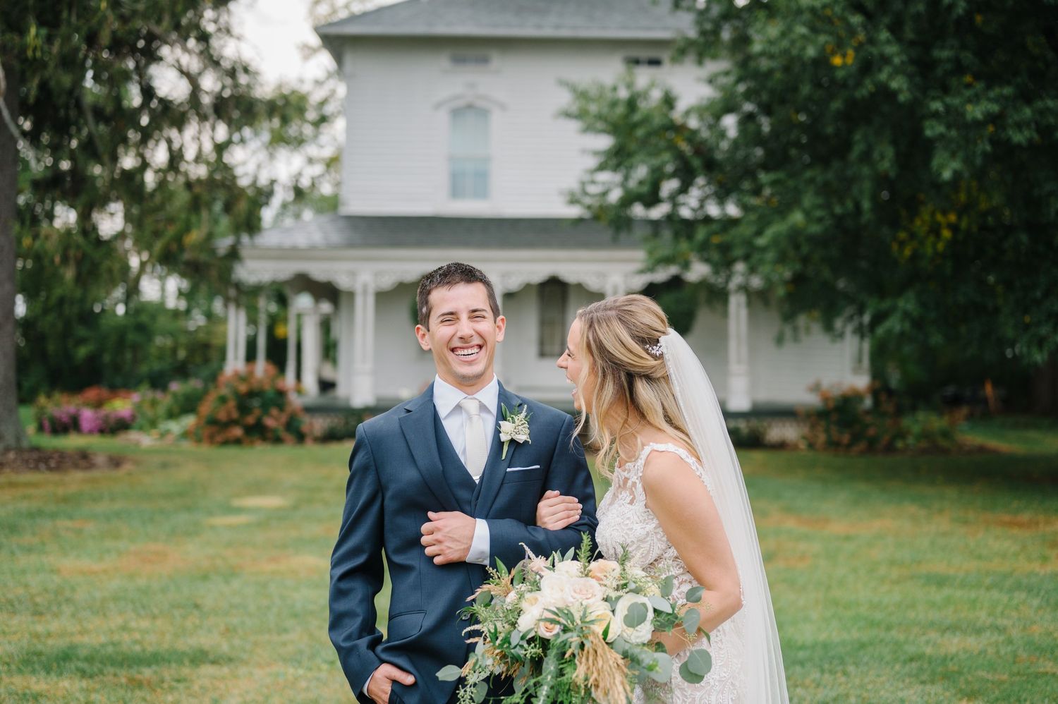 Newlywed couple shares a joyful moment together on a lawn in front of a white farmhouse on their wedding day.