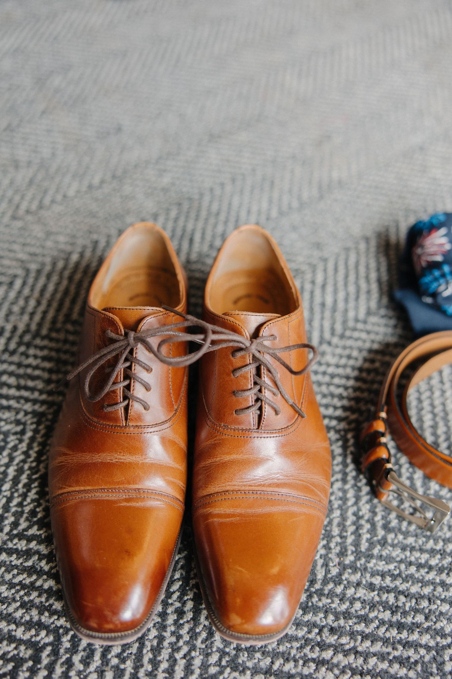 A pair of polished brown leather oxford dress shoes with laces rests on a gray herringbone carpet.