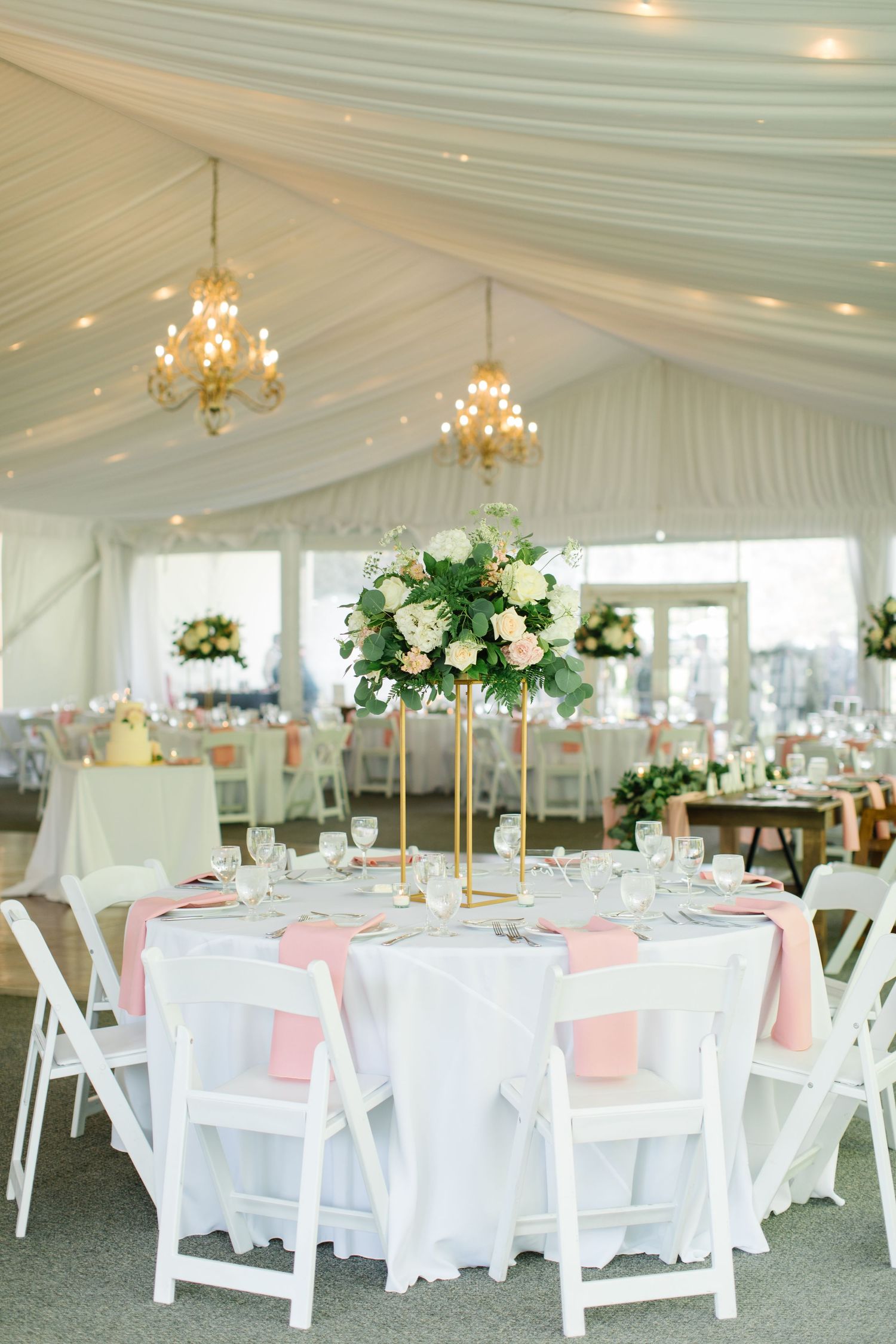 Elegant wedding reception tent with white draping, crystal chandeliers, pink napkins and tall floral centerpieces on round tables.