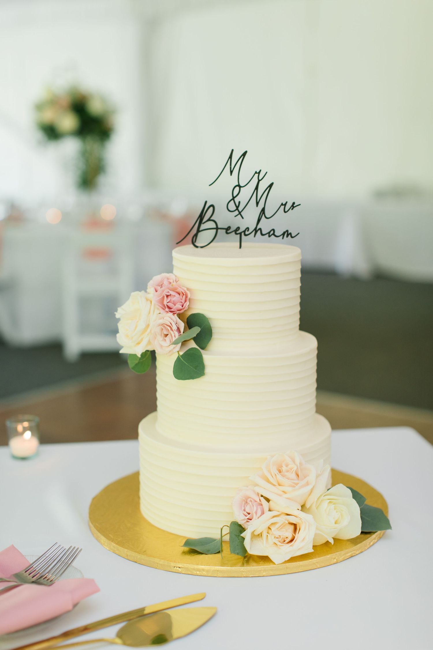 Elegant three-tiered white wedding cake with pink and cream roses on a gold cake stand with custom name topper.