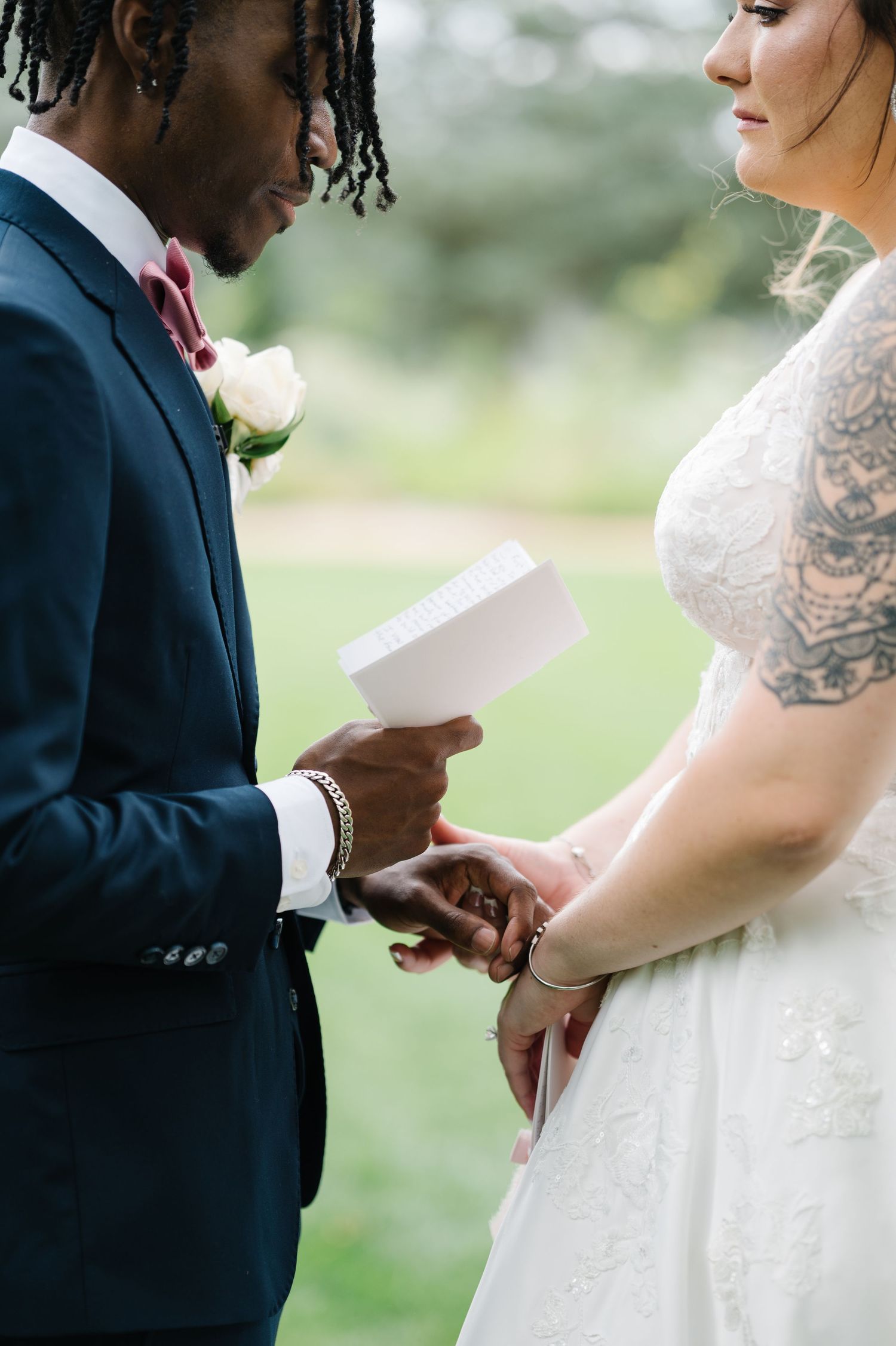 A couple exchanges vows during an intimate outdoor wedding ceremony, with the groom holding papers and the bride in lace.