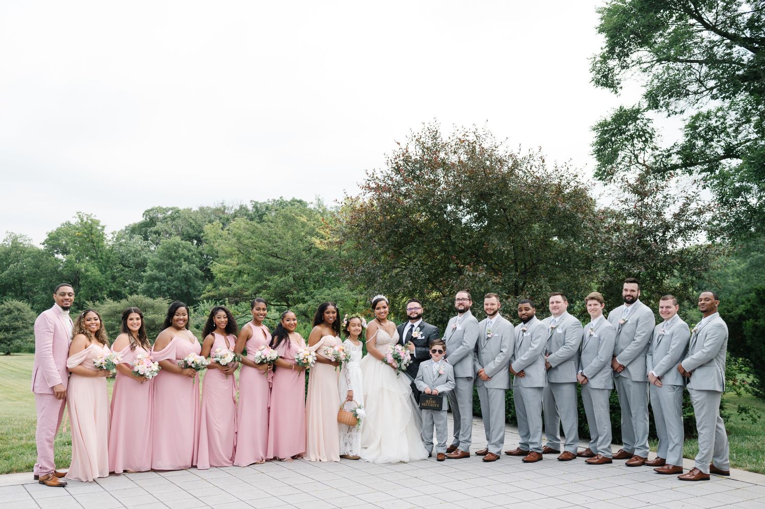 Wedding party in pink and gray attire poses outdoors in a natural setting with trees in the background.
