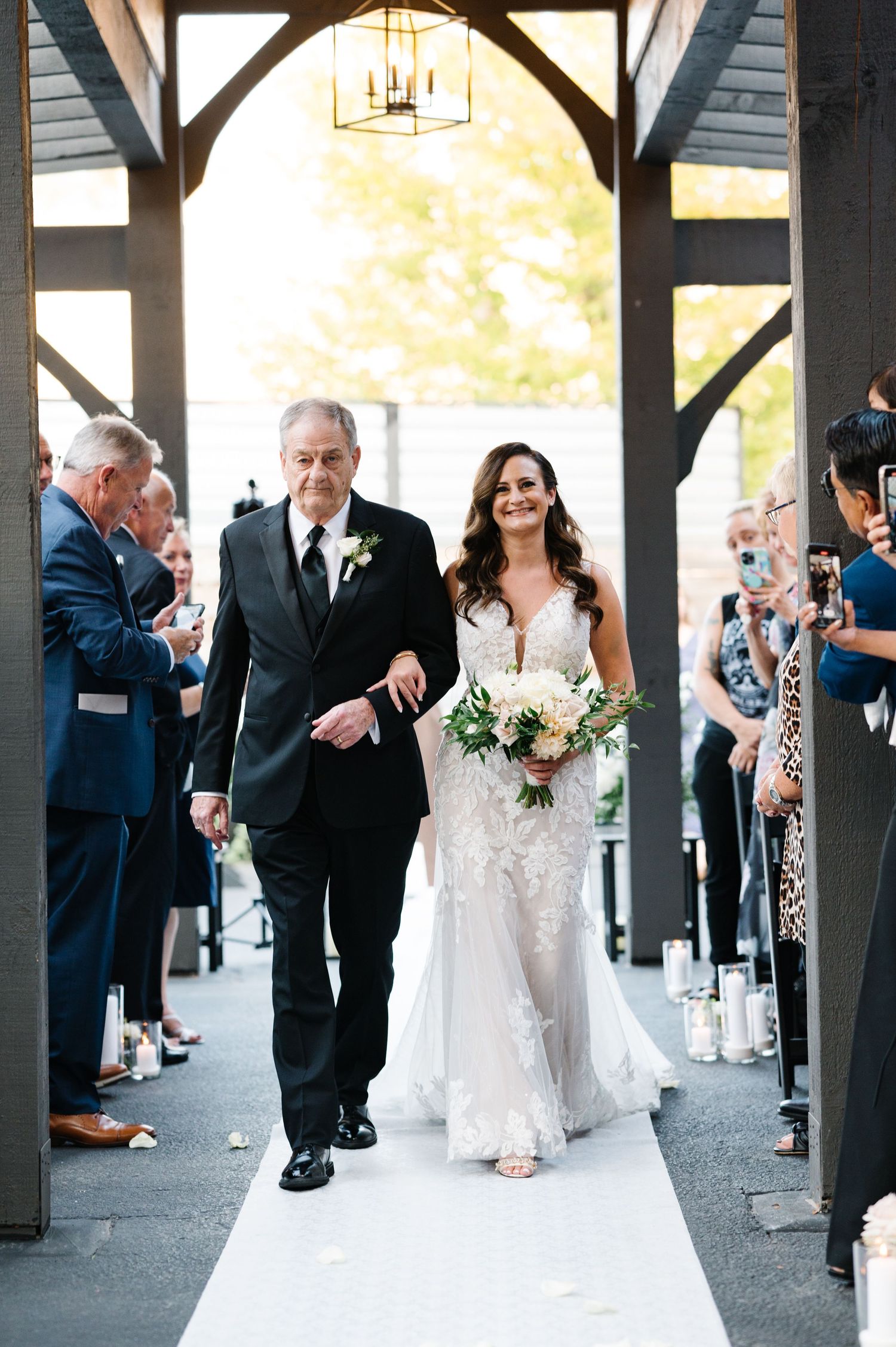 Beautiful wedding ceremony moment as bride walks down white aisle under rustic wooden arch with guests watching.
