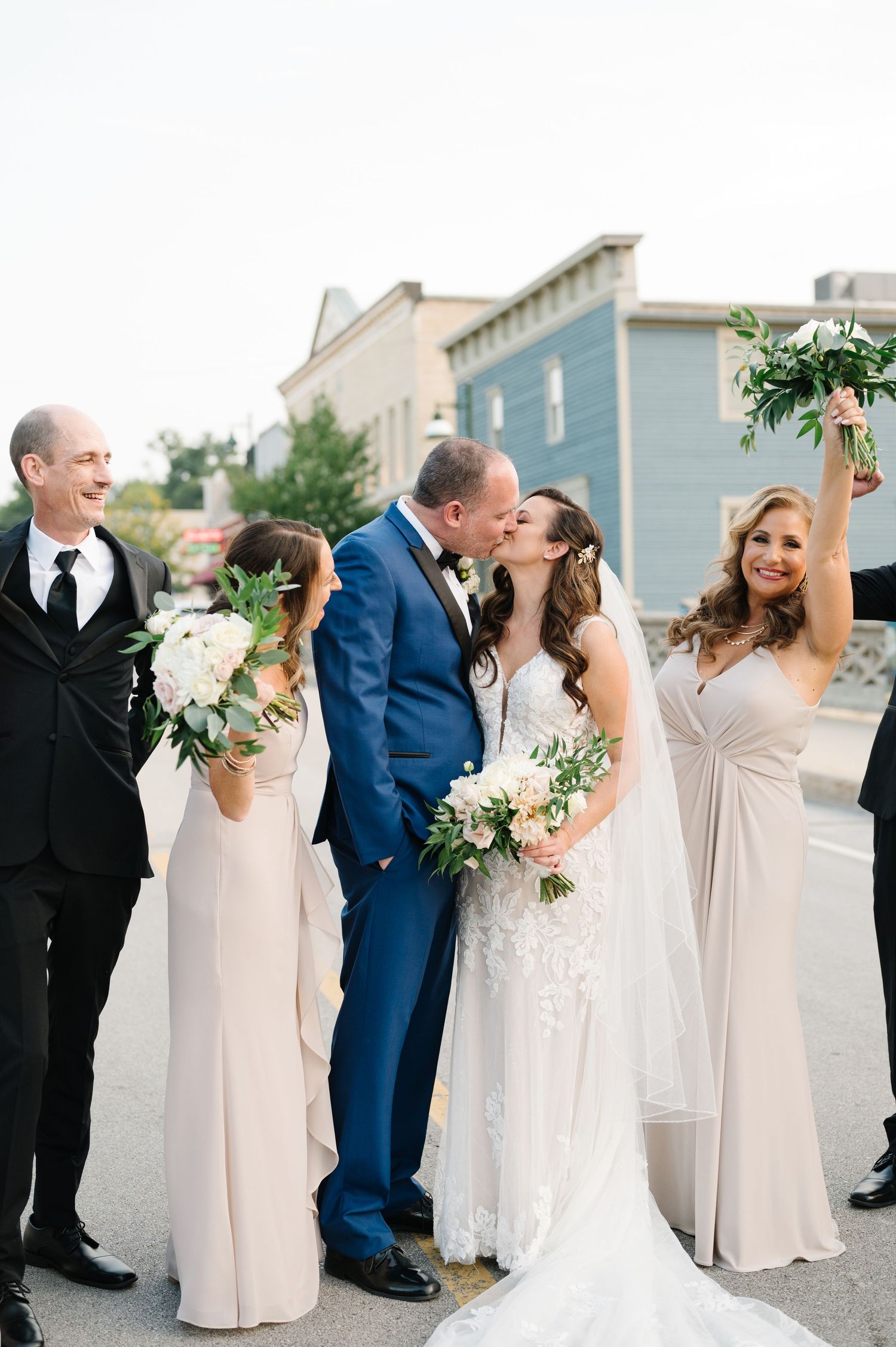 Wedding party in formal attire with bride in white dress and bridesmaids in blush gowns pose on a city street.