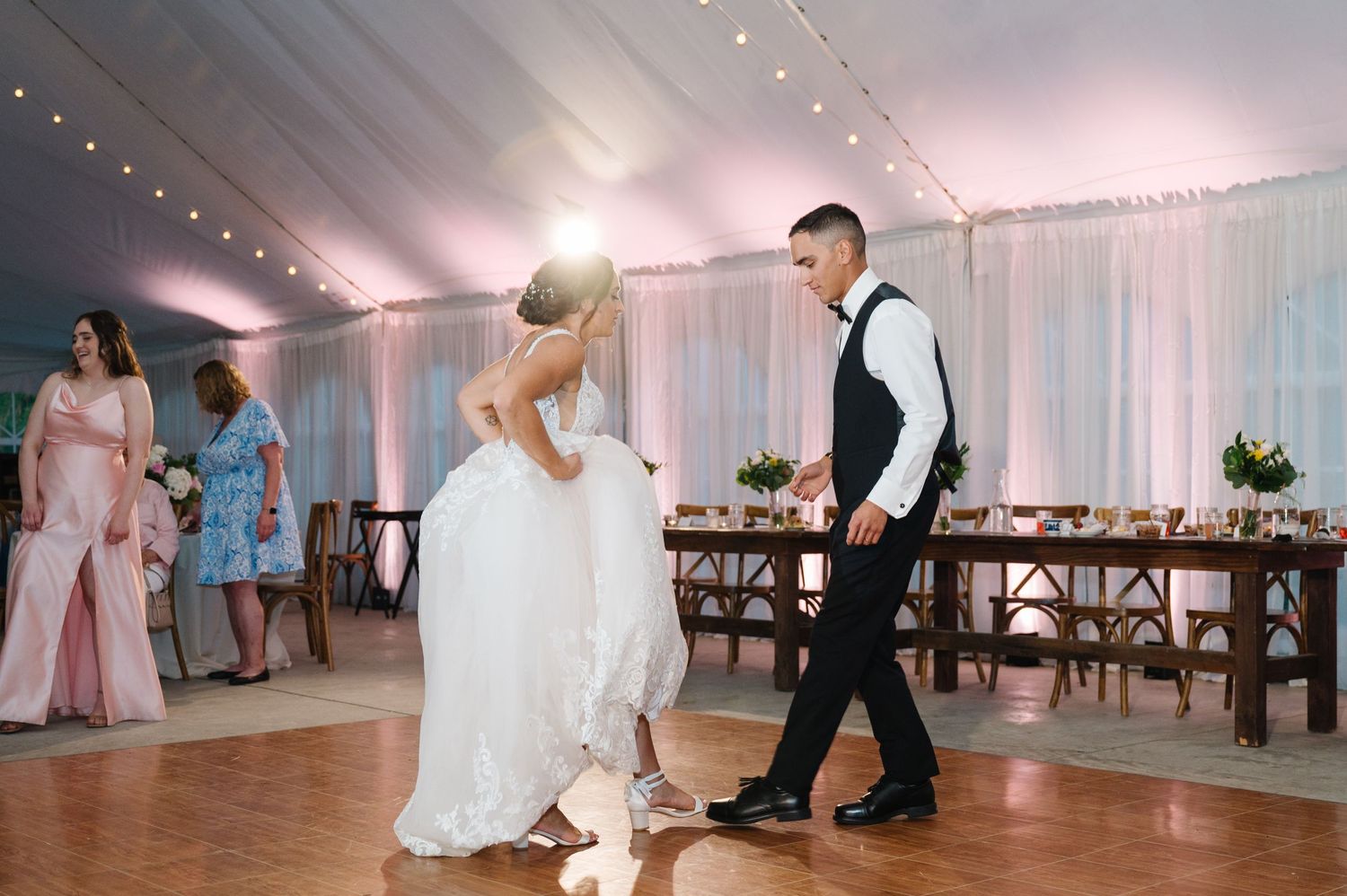 Newlyweds share their first dance together in an elegant white-draped reception tent with string lights overhead.