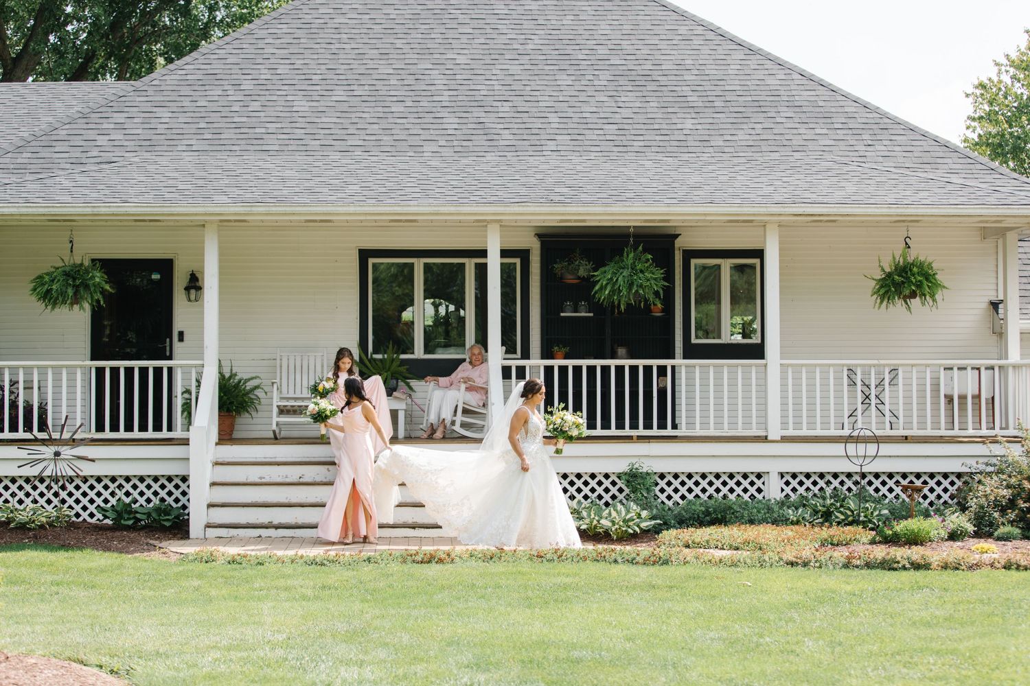 Two women in flowing dresses stand on a white porch of a charming farmhouse with hanging plants and lattice details.