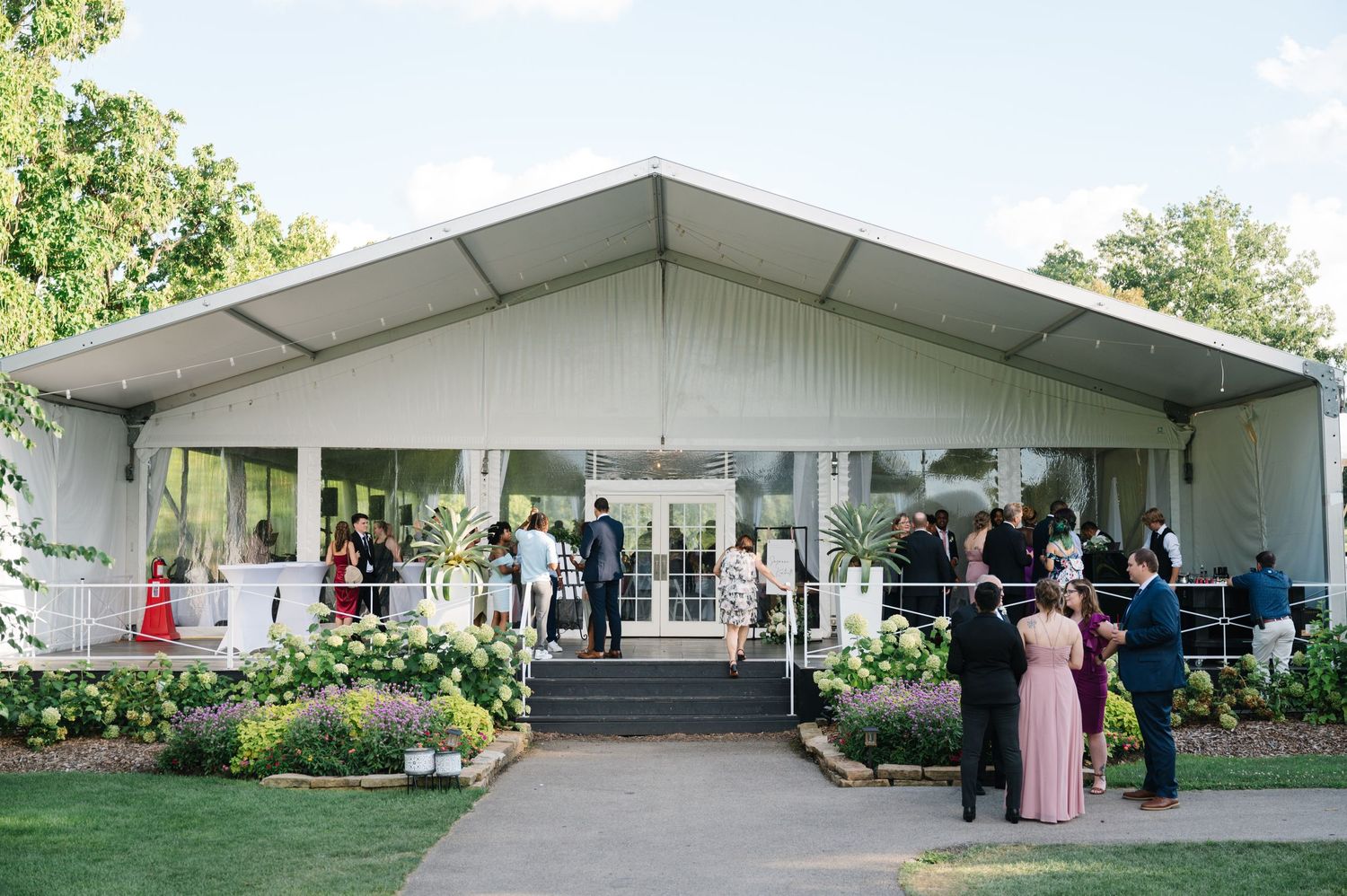Event guests gather outside a white tent pavilion with landscaped gardens and flower arrangements at the entrance.