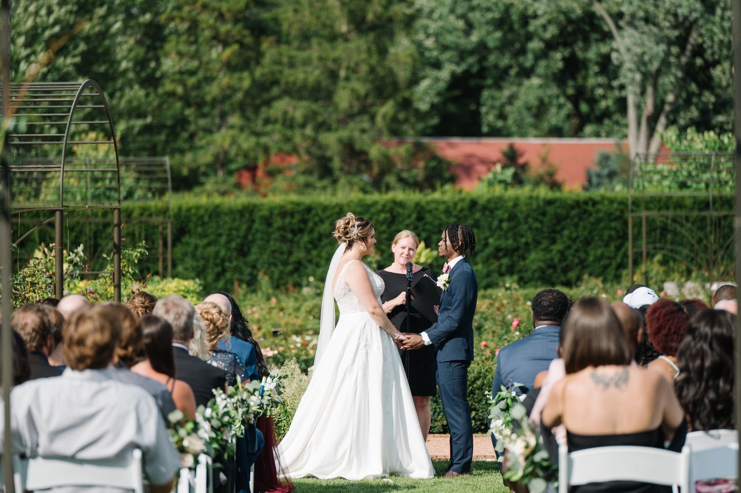 An outdoor garden wedding ceremony takes place with an officiant and couple surrounded by guests and manicured hedges.