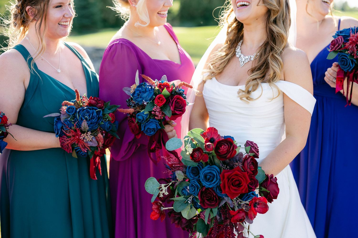Wedding party in jewel-toned dresses holding vibrant bouquets with red and blue flowers on sunny outdoor lawn.
