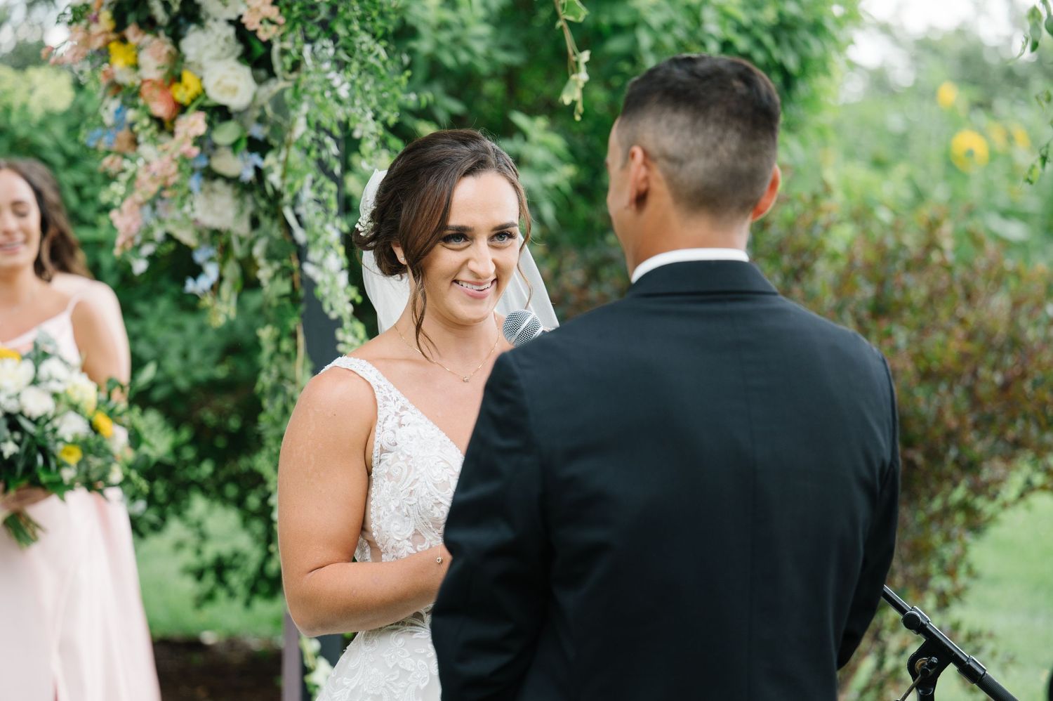 Outdoor wedding ceremony with a couple exchanging vows under a lush green floral arch and natural setting.