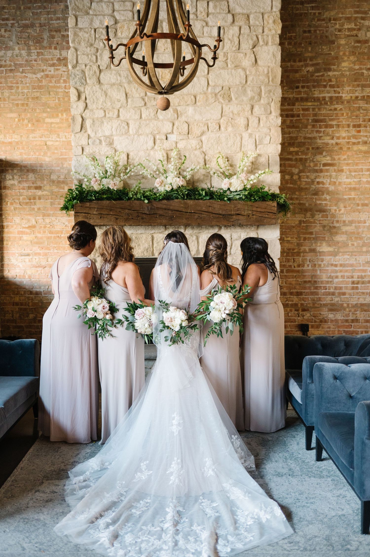 Bridesmaids in pale dresses gather with bride in lace gown against exposed brick wall beneath rustic chandelier.