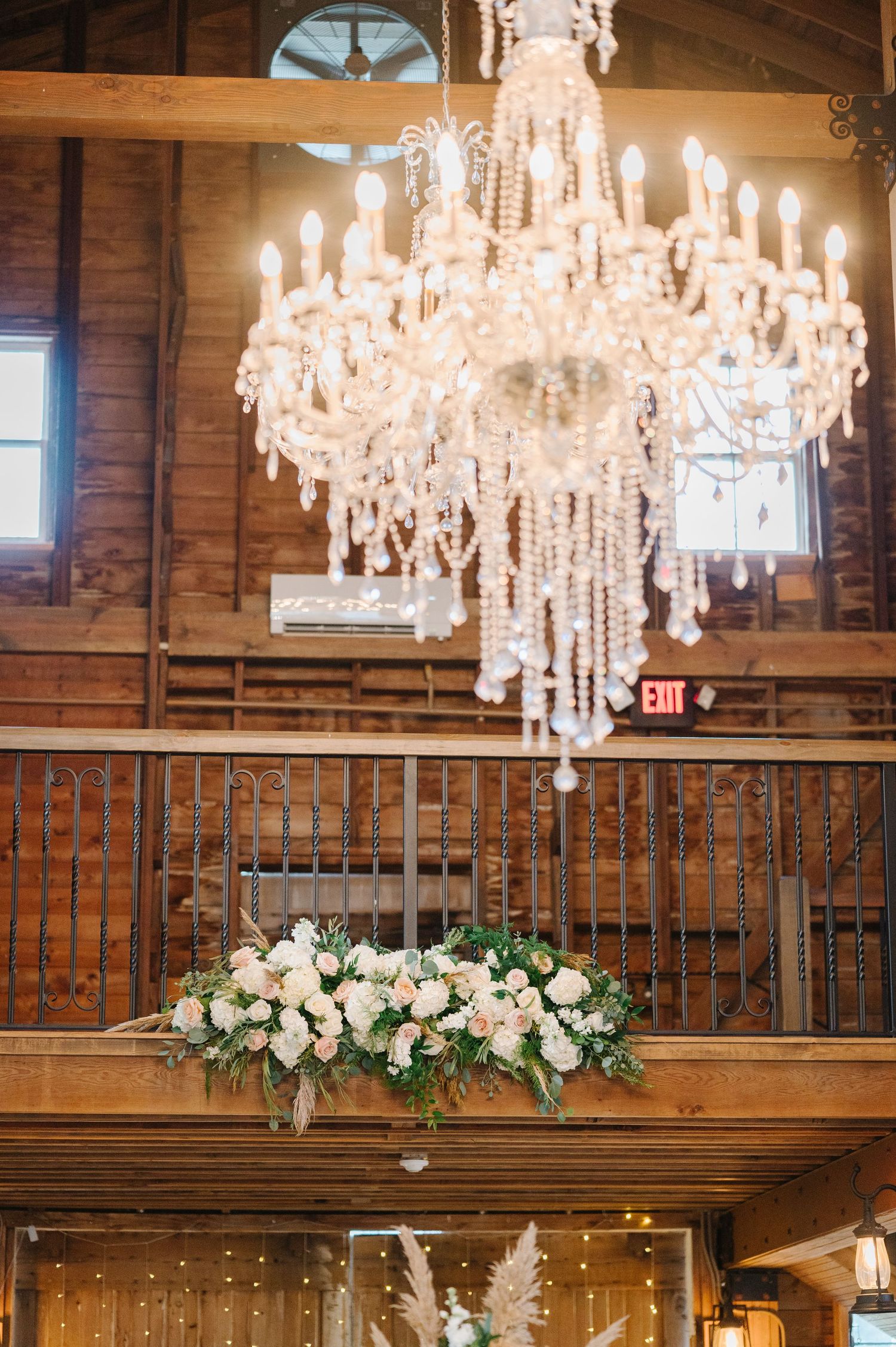 An elegant crystal chandelier illuminates a rustic wooden interior with a floral arrangement on a balcony railing below.