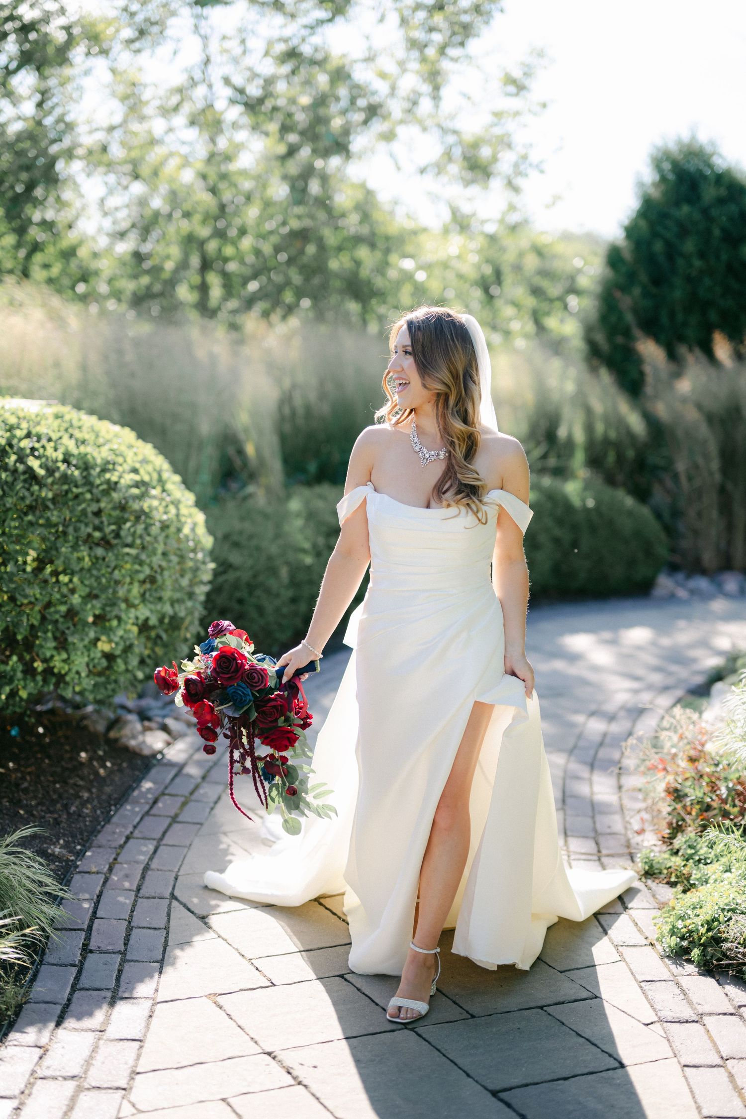 Elegant bride in flowing off-shoulder white dress holds vibrant red bouquet while walking along garden path on sunny day.