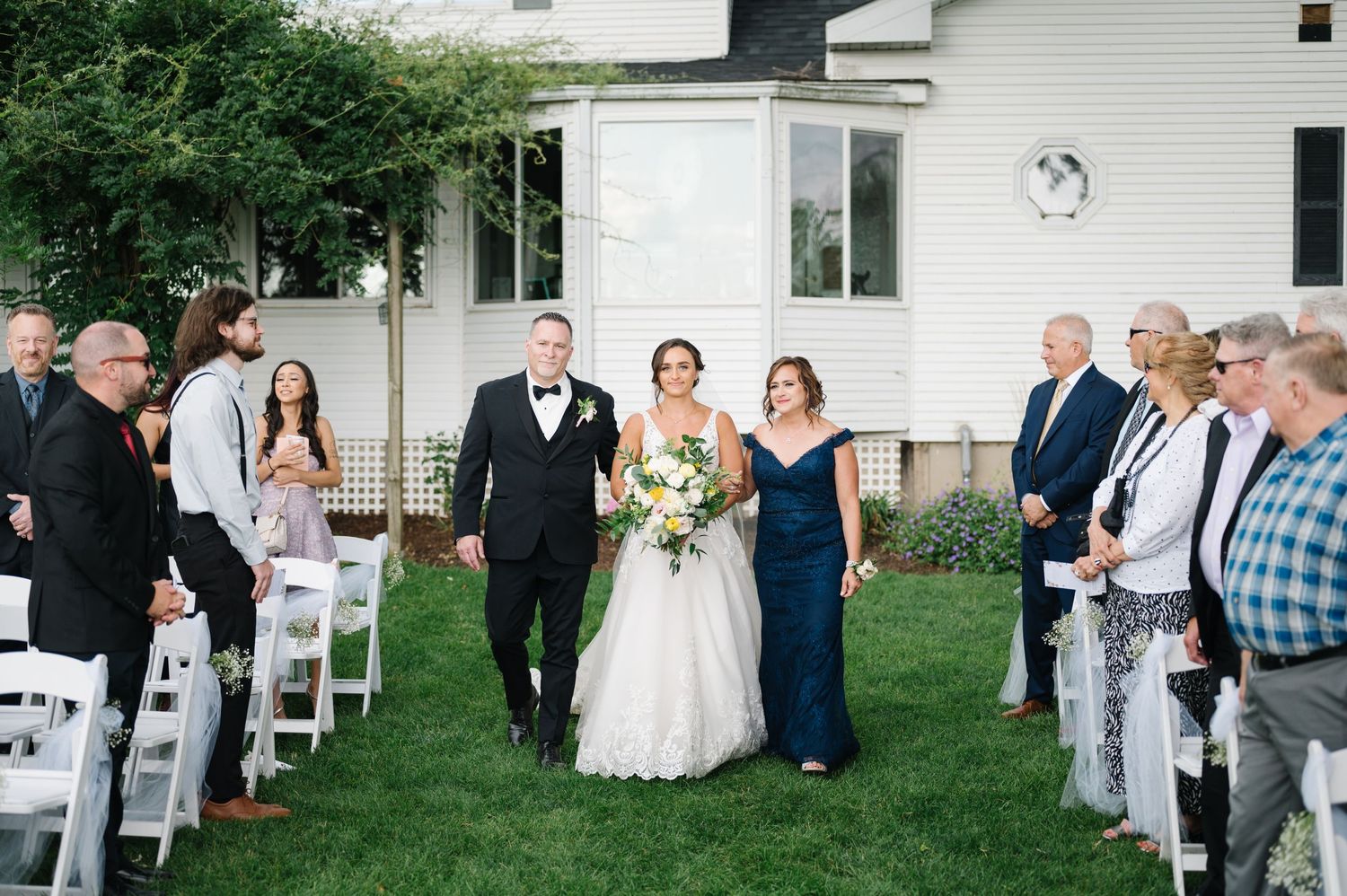 An outdoor wedding ceremony on a green lawn beside a white house with guests seated in white folding chairs.