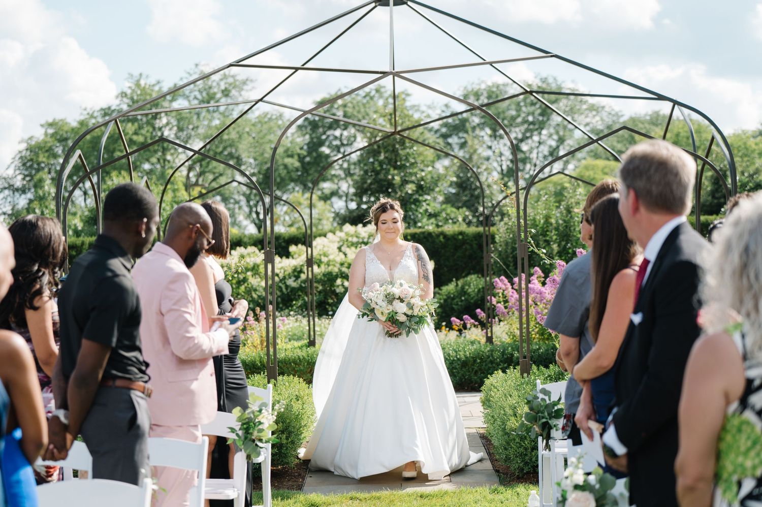 An outdoor wedding ceremony takes place under an elegant gazebo surrounded by lush greenery and flowering gardens.