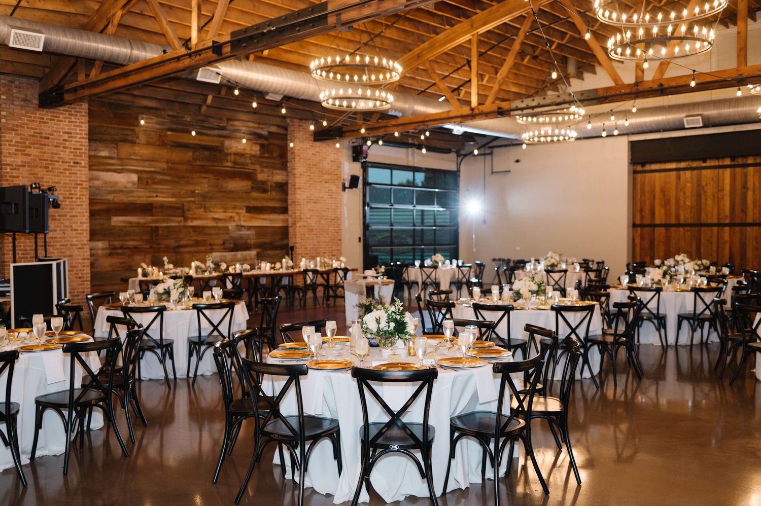 Modern rustic wedding venue with wooden walls, industrial chandeliers, and black cross-back chairs around white-clothed tables.