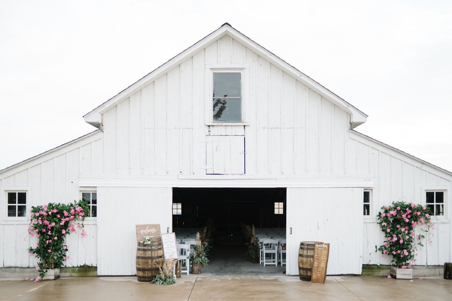 A white rustic barn with climbing roses features open doors and wooden barrels at the entrance for a romantic venue setup.