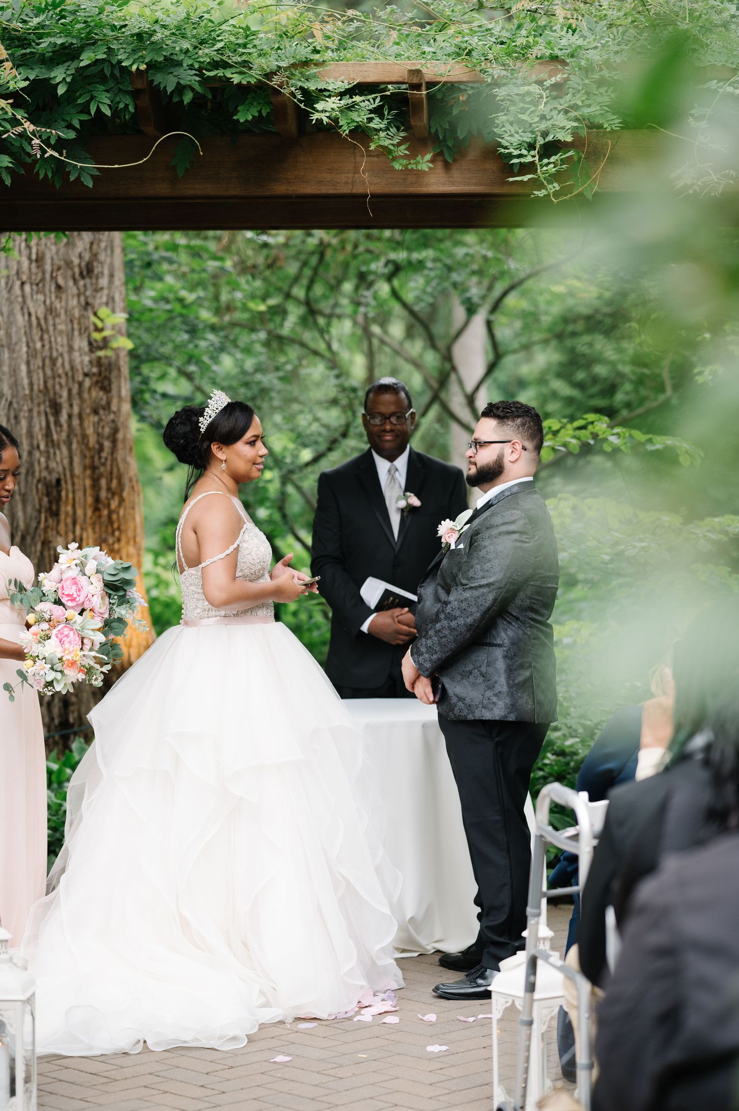 An outdoor wedding ceremony taking place under a wooden pergola surrounded by lush greenery and decorated with pink flowers.