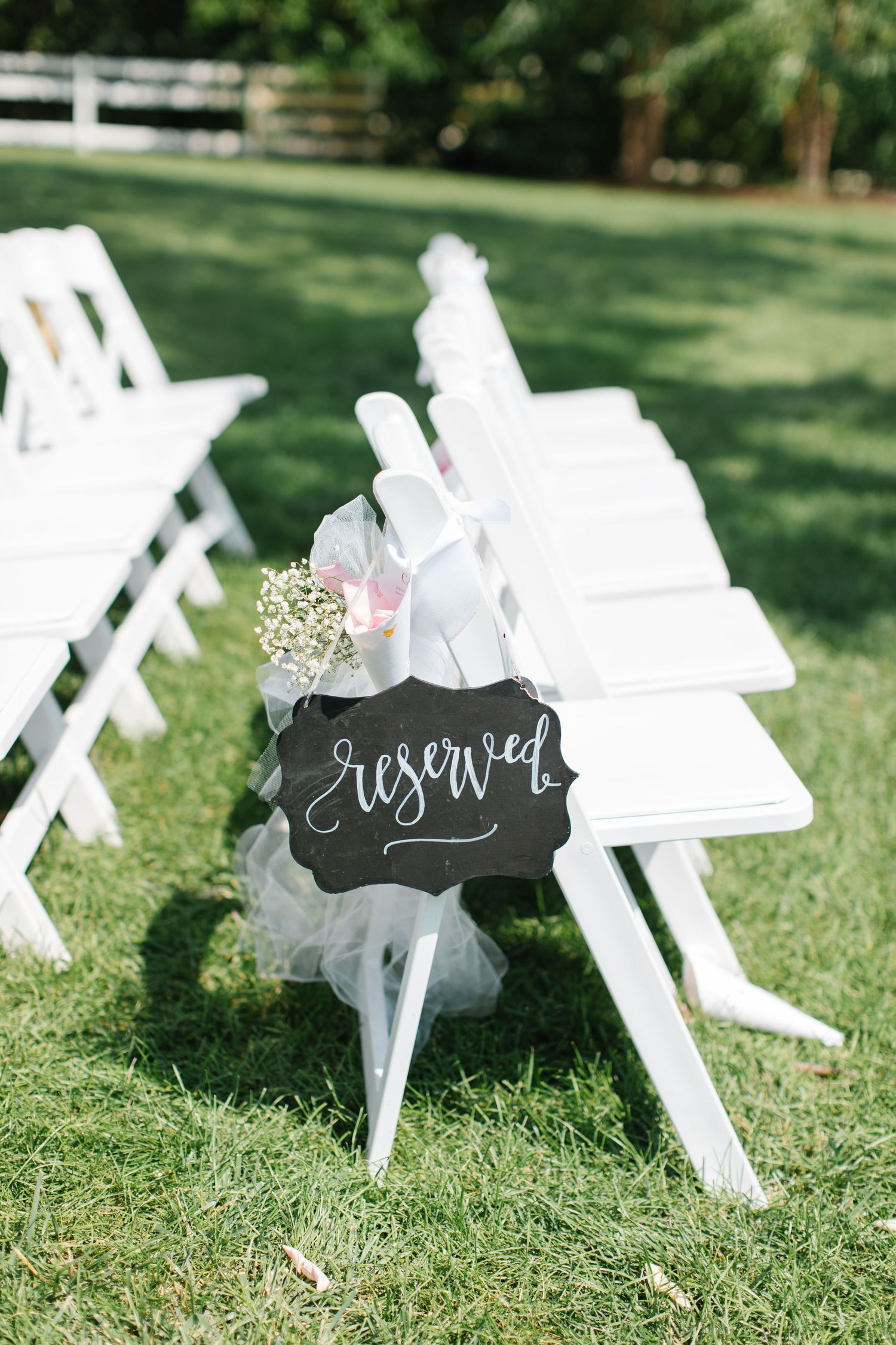White wooden wedding chairs with a chalkboard 'reserved' sign and floral decoration in an outdoor garden setting.