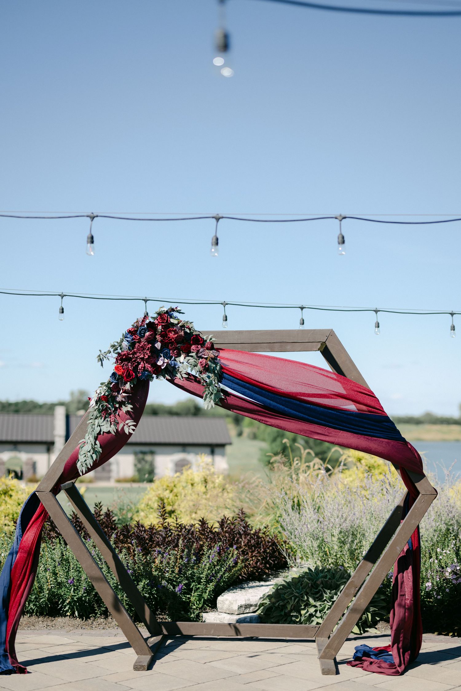 A wooden hexagonal wedding arch decorated with burgundy fabric and flowers under string lights against a blue sky.