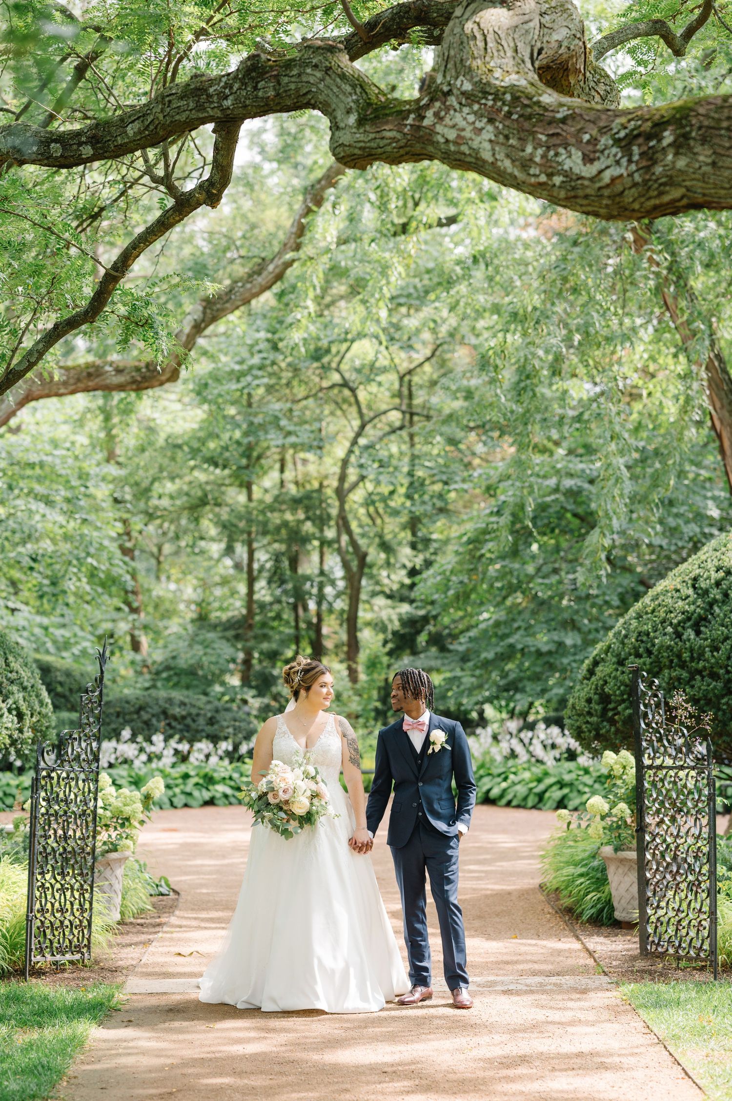 A couple stands together on a romantic garden path beneath towering trees and an ornate metal archway.