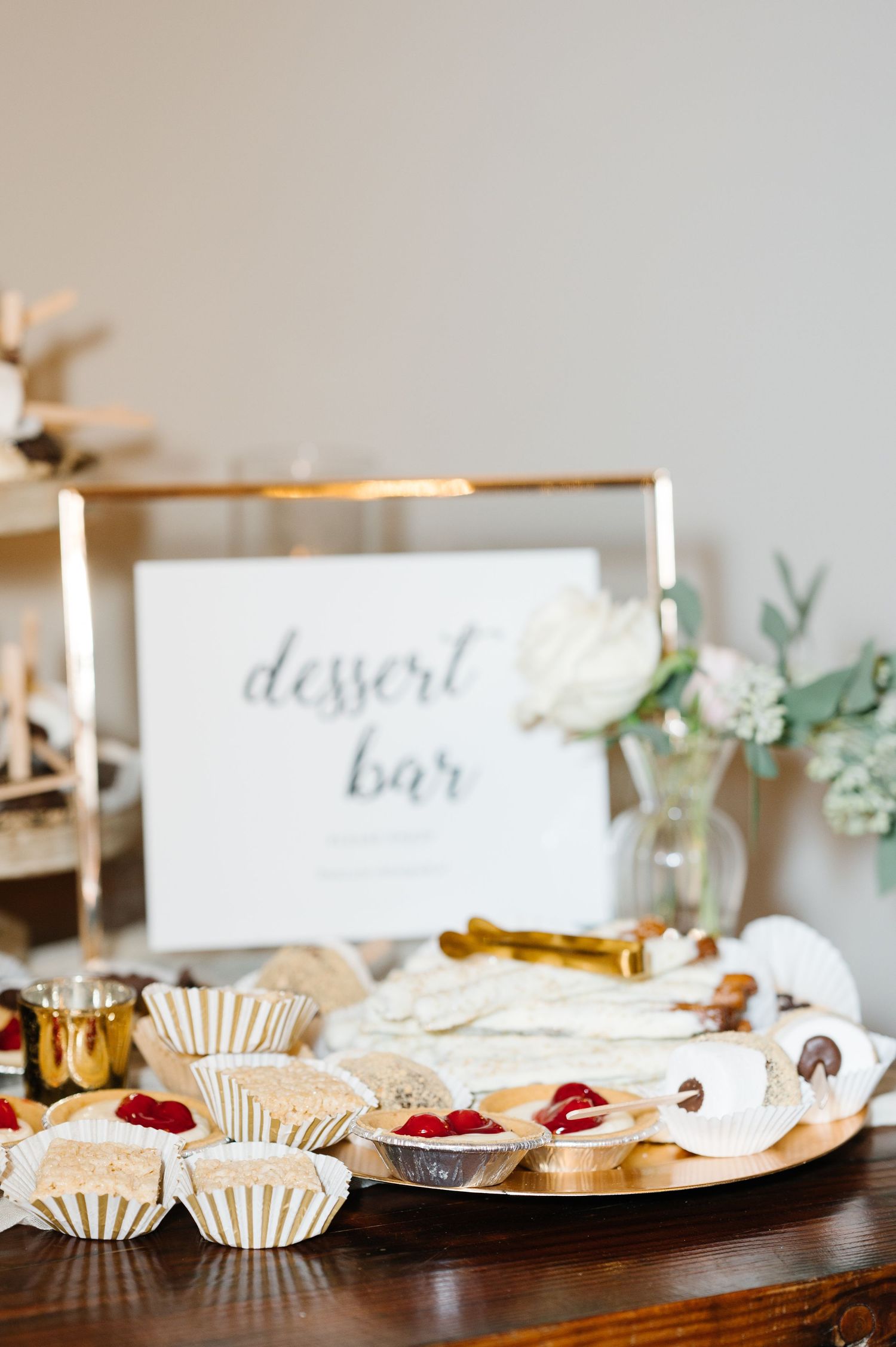 A rustic dessert bar table setup with white calligraphy signage, paper cupcake liners, and fresh floral accents.