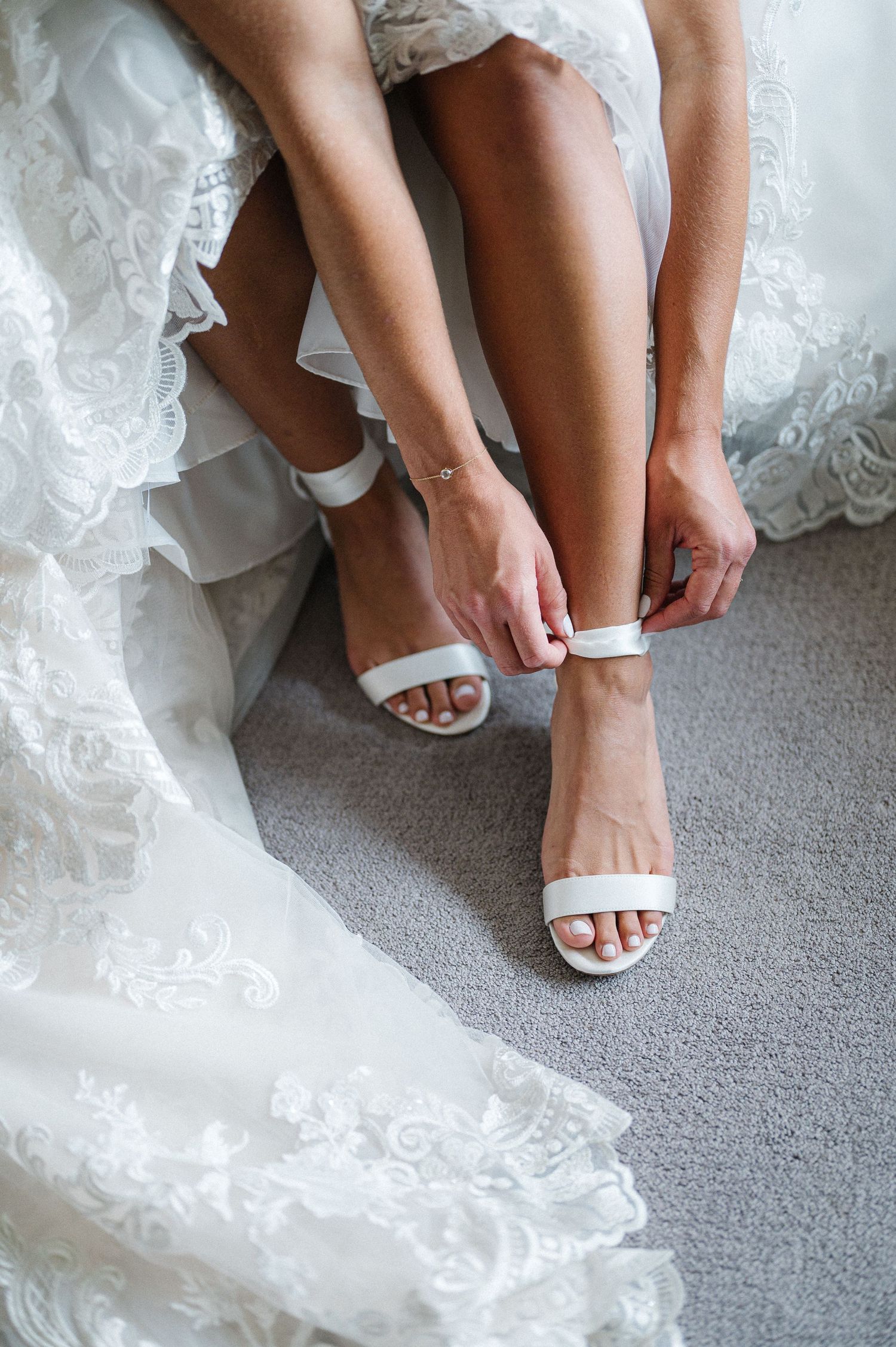 Hands fastening delicate white ankle strap sandals with lace wedding dress details visible on grey carpet.