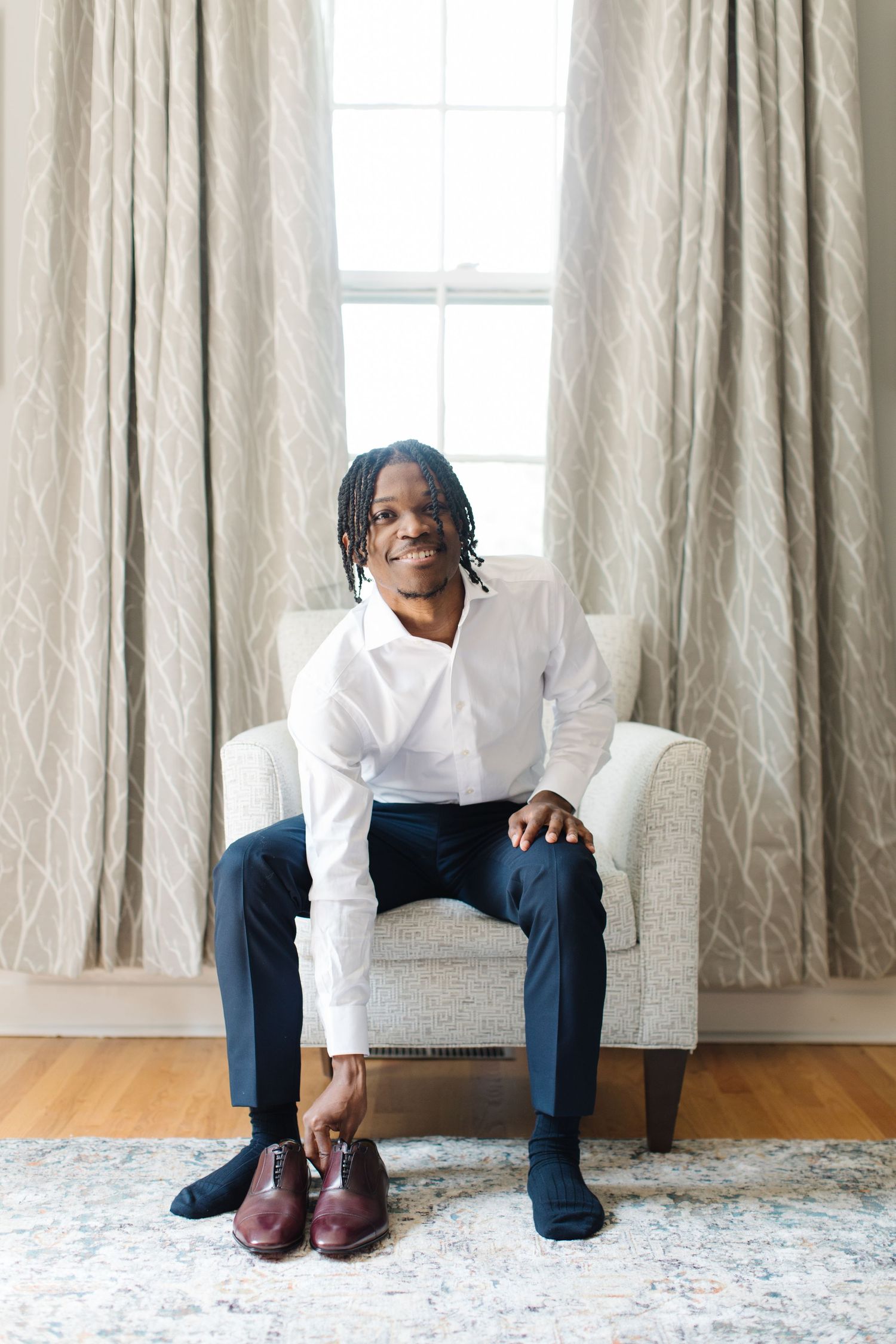 A person in a white sweater and navy pants sits on a grey armchair, smiling warmly by a curtained window.
