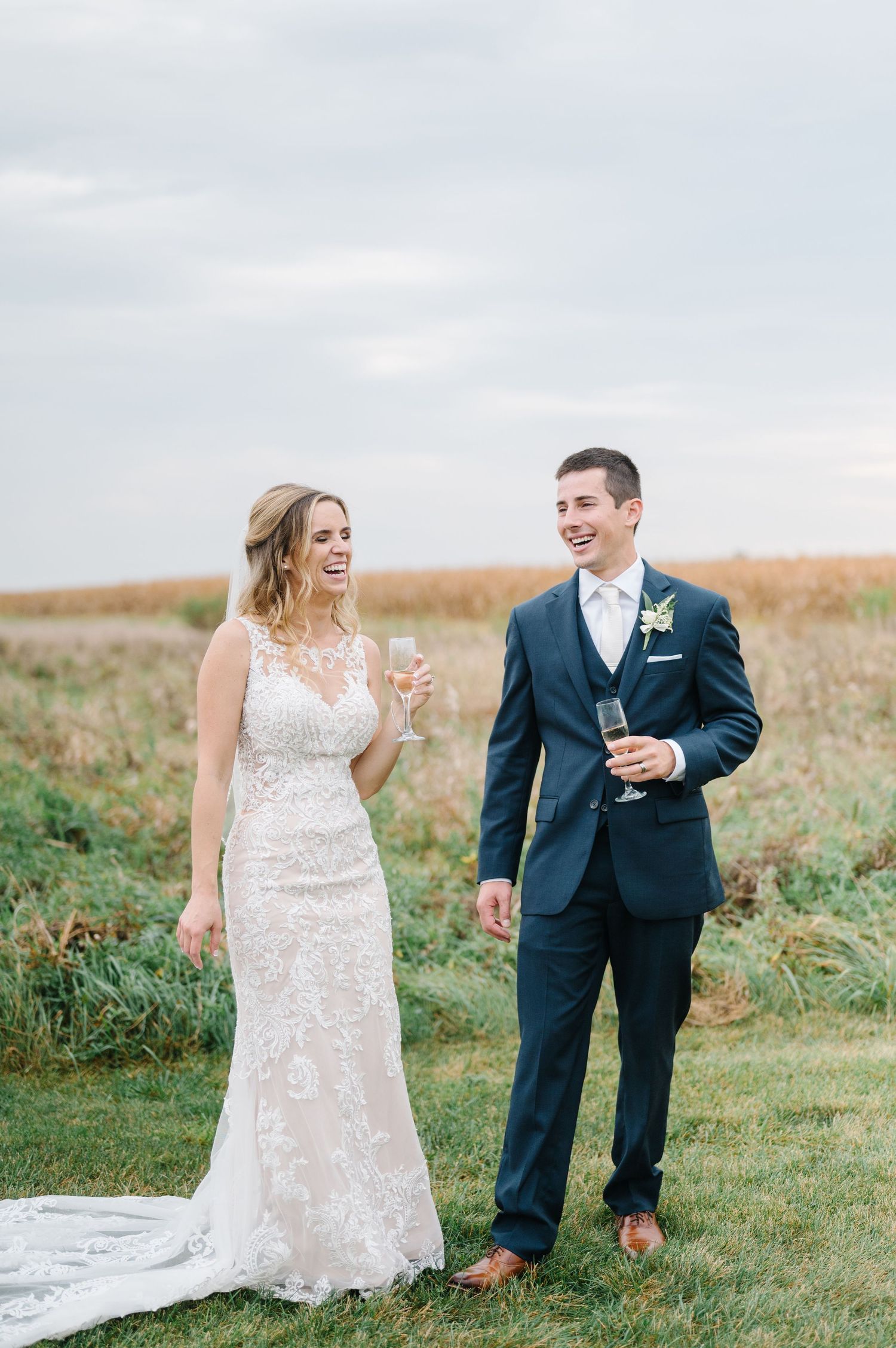 A bride in a lace gown and groom in navy suit share champagne together on a hilltop during their outdoor wedding.