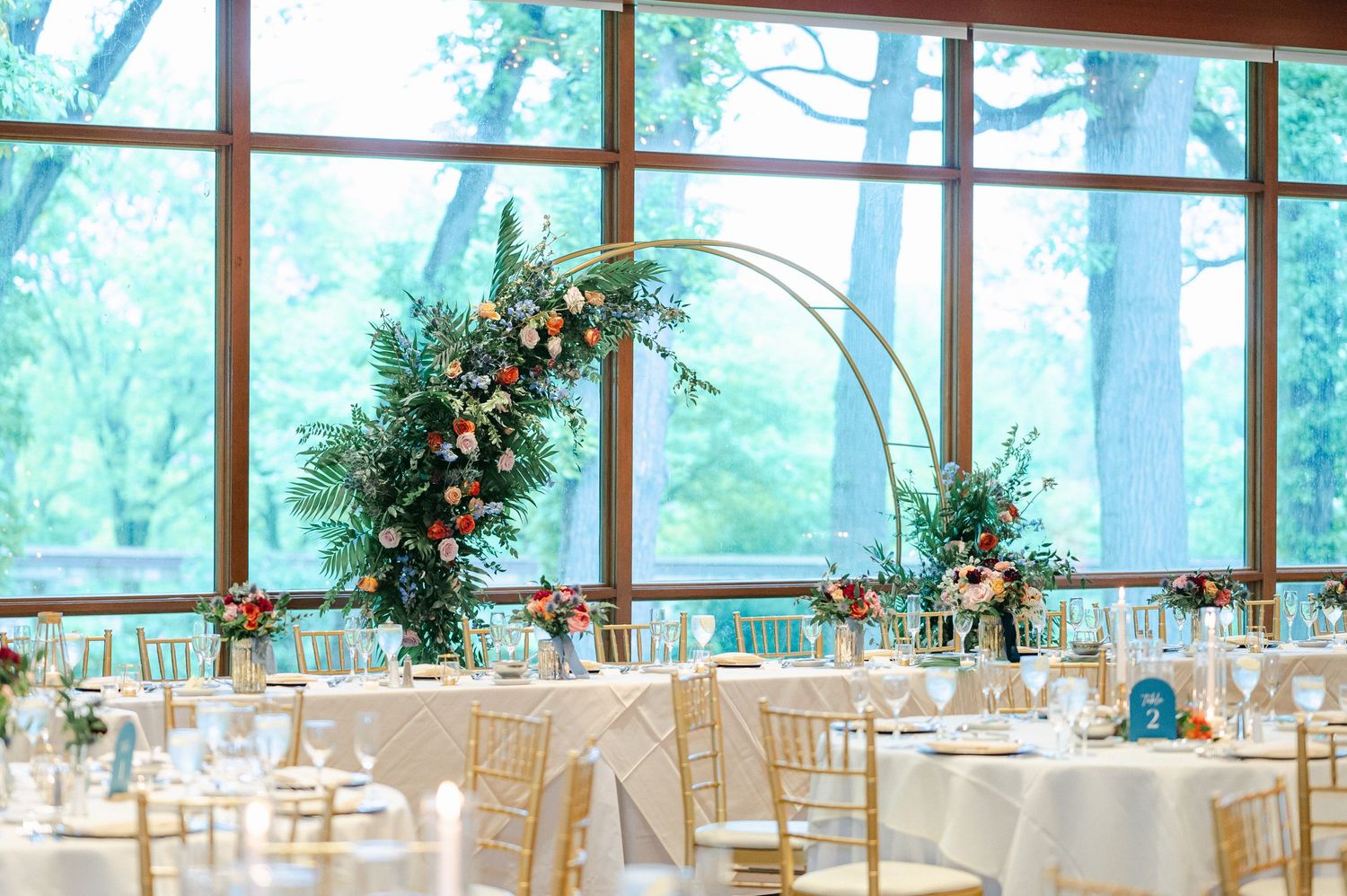 A light-filled wedding reception venue with white tables, chiavari chairs, and tall floral arch decorations against forest views.