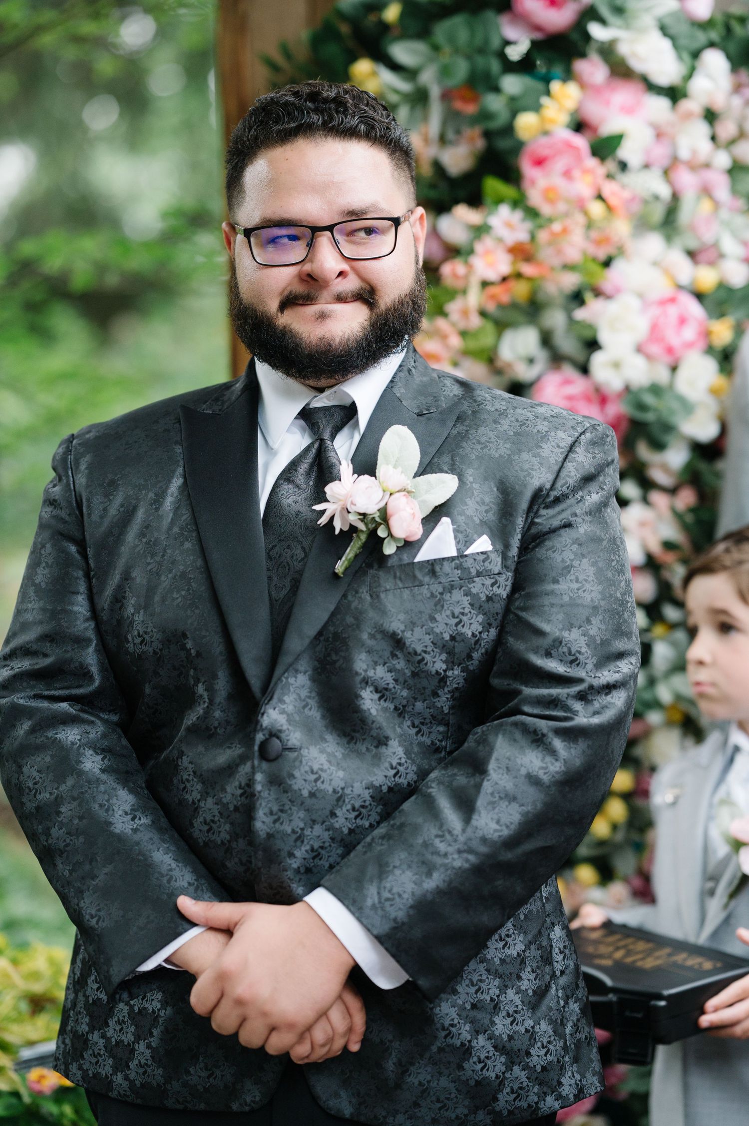 A groom wearing a charcoal patterned suit and glasses stands at the wedding altar with floral arrangements in background.