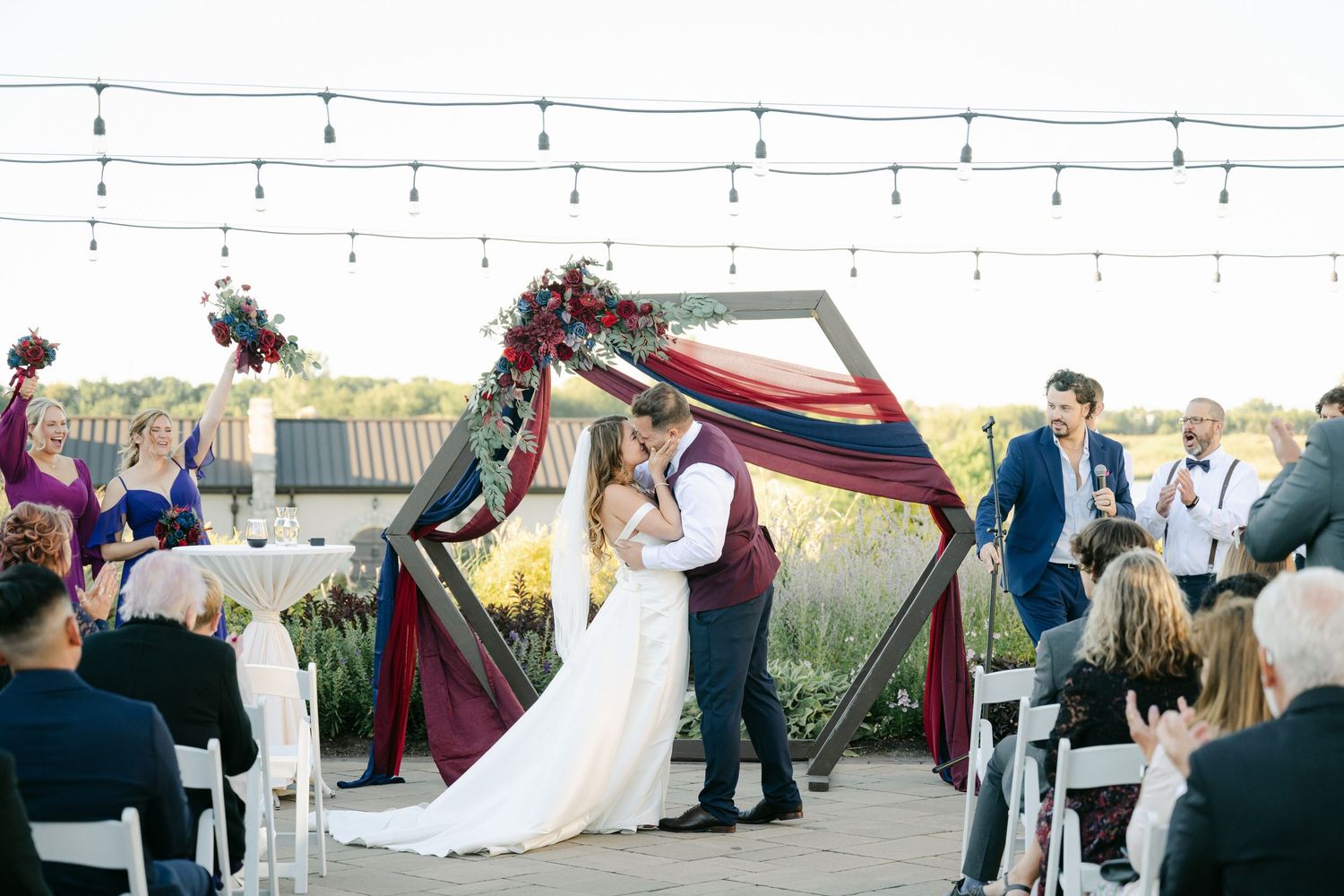 An outdoor wedding ceremony under a decorated hexagonal arch with string lights and colorful draping overhead.