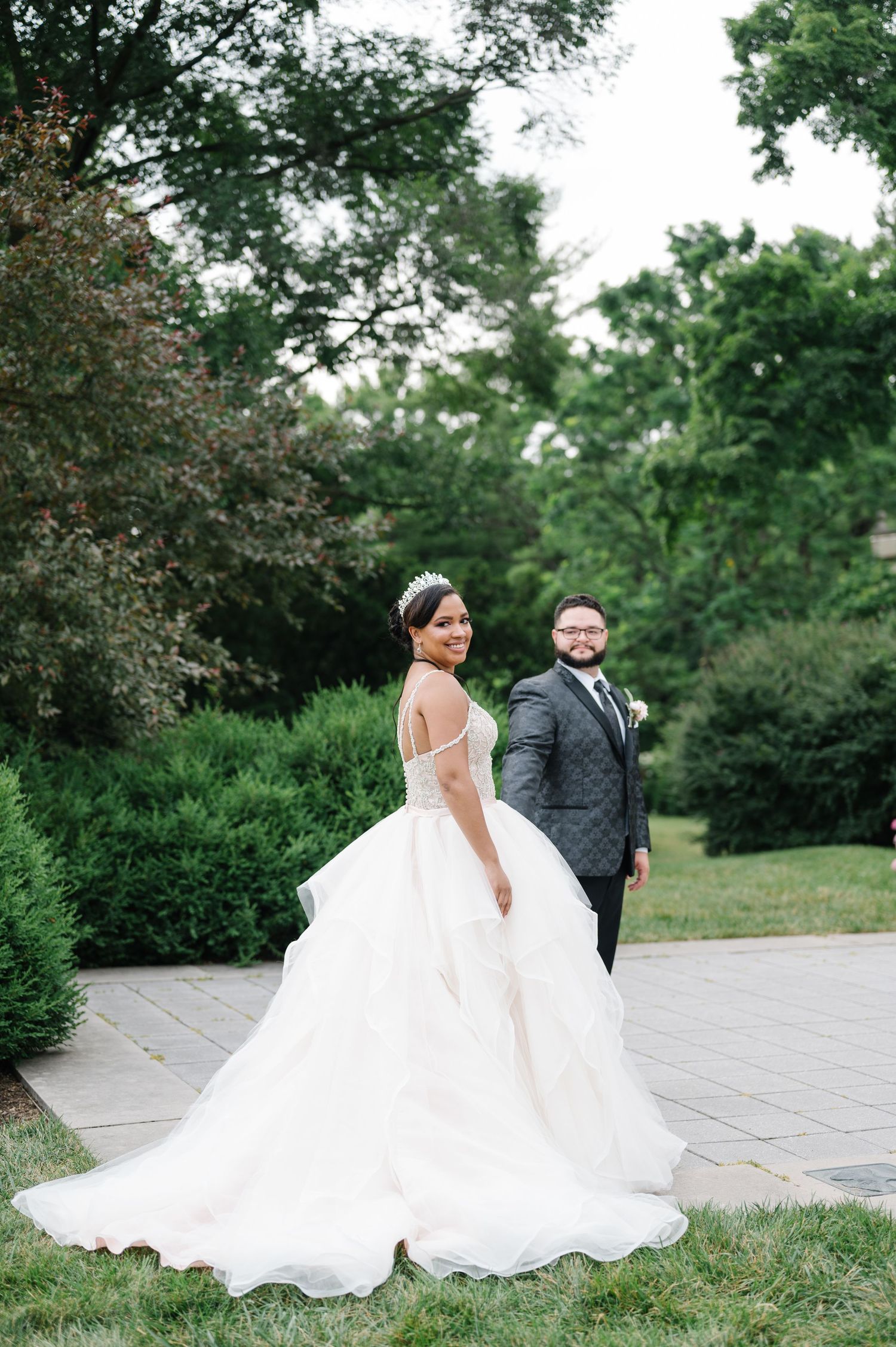 A wedding couple stands together outdoors on a stone path surrounded by lush green gardens and trees.