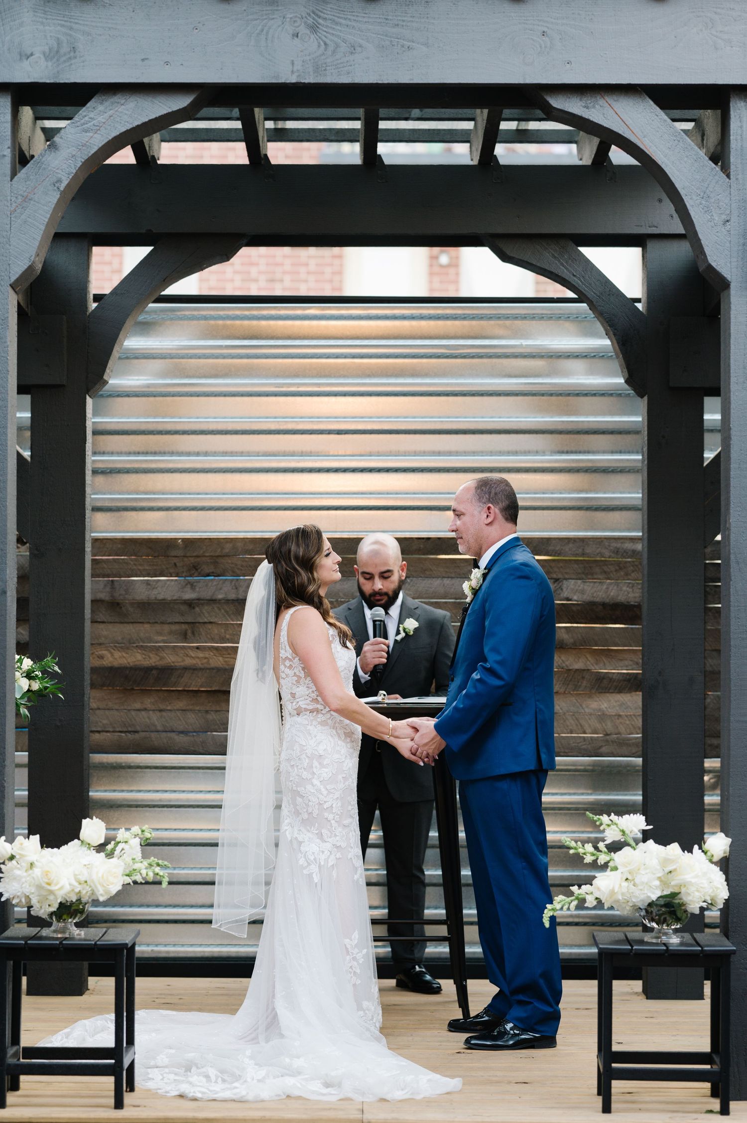 A wedding ceremony takes place under a wooden archway with metal details and white floral decorations.
