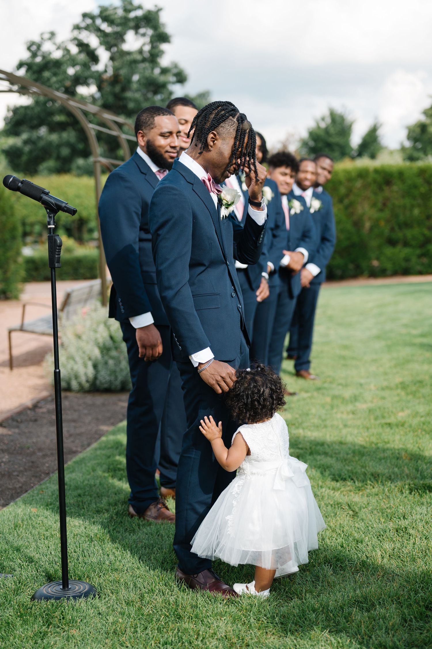 A groomsmen line stands at an outdoor wedding ceremony while a small child in a white dress embraces one of them on a lawn.