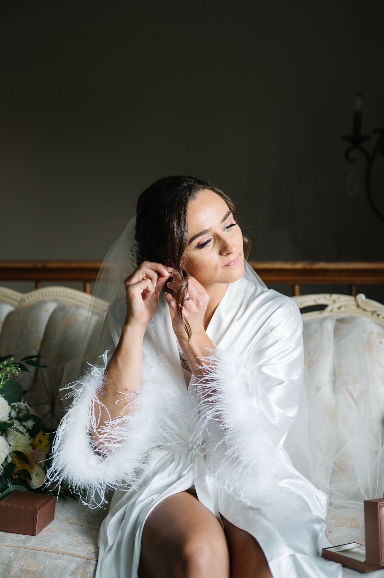 A bride in a white silk robe with feather trim poses gracefully on an elegant vintage sofa during her wedding preparation.