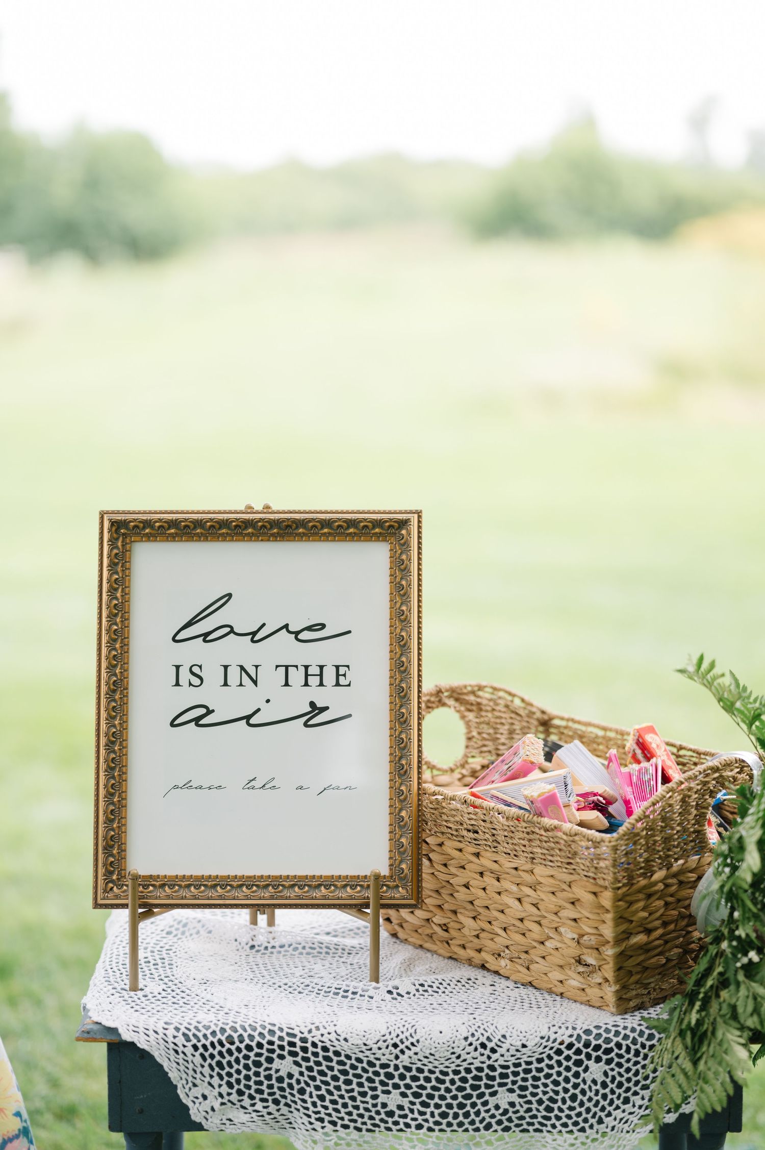 Wedding reception decor with framed 'Love is in the air' sign and woven basket on lace-covered table outdoors.