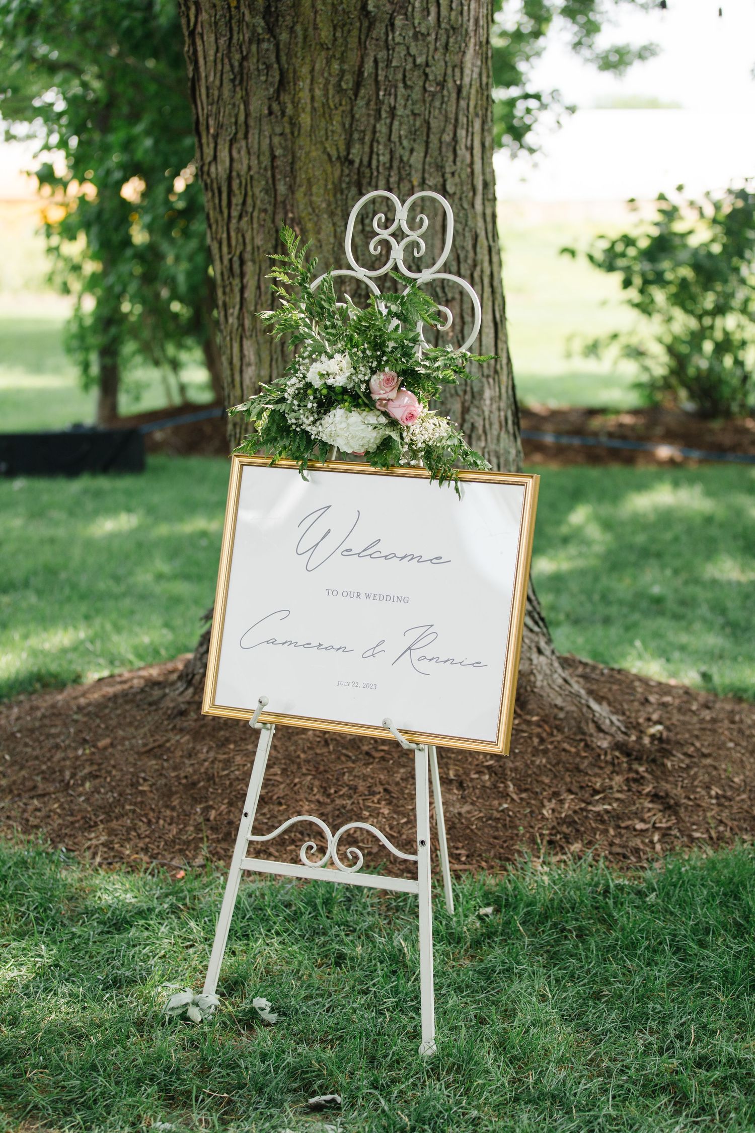 Elegant wedding welcome sign with floral arrangement displayed on white metal easel in an outdoor garden setting. - Heritage Prairie Farm