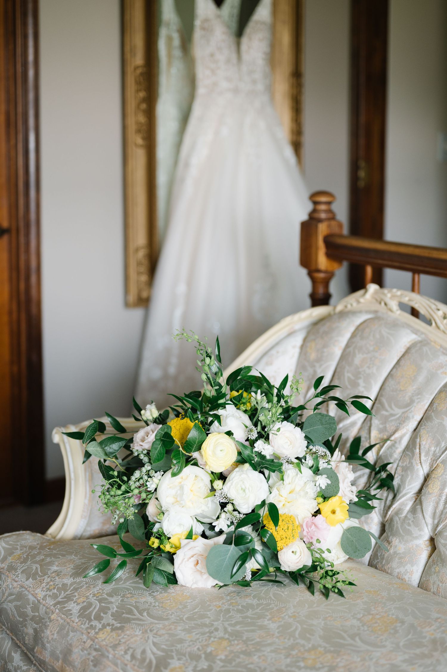 Wedding bouquet with white and yellow flowers rests on vintage chair with wedding dress hanging in background.