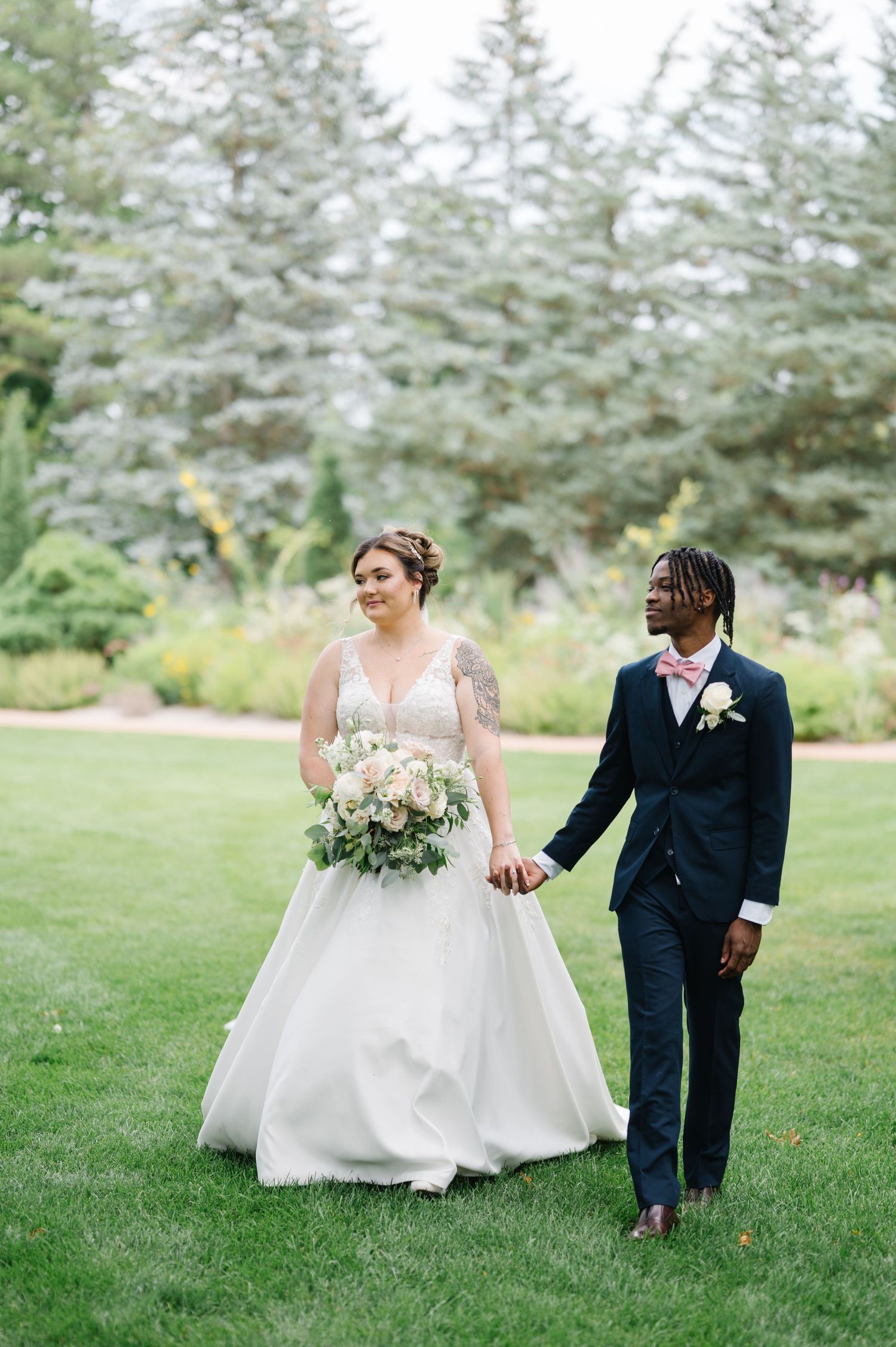 Two newlyweds walking hand in hand on lush green lawn with evergreen trees in background.