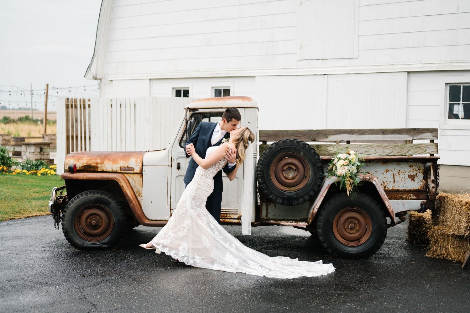 A rustic wedding photo by an old vintage Jeep with hay bales at a white barn on a rainy day. - Ashley Farm in Yorkville, IL
