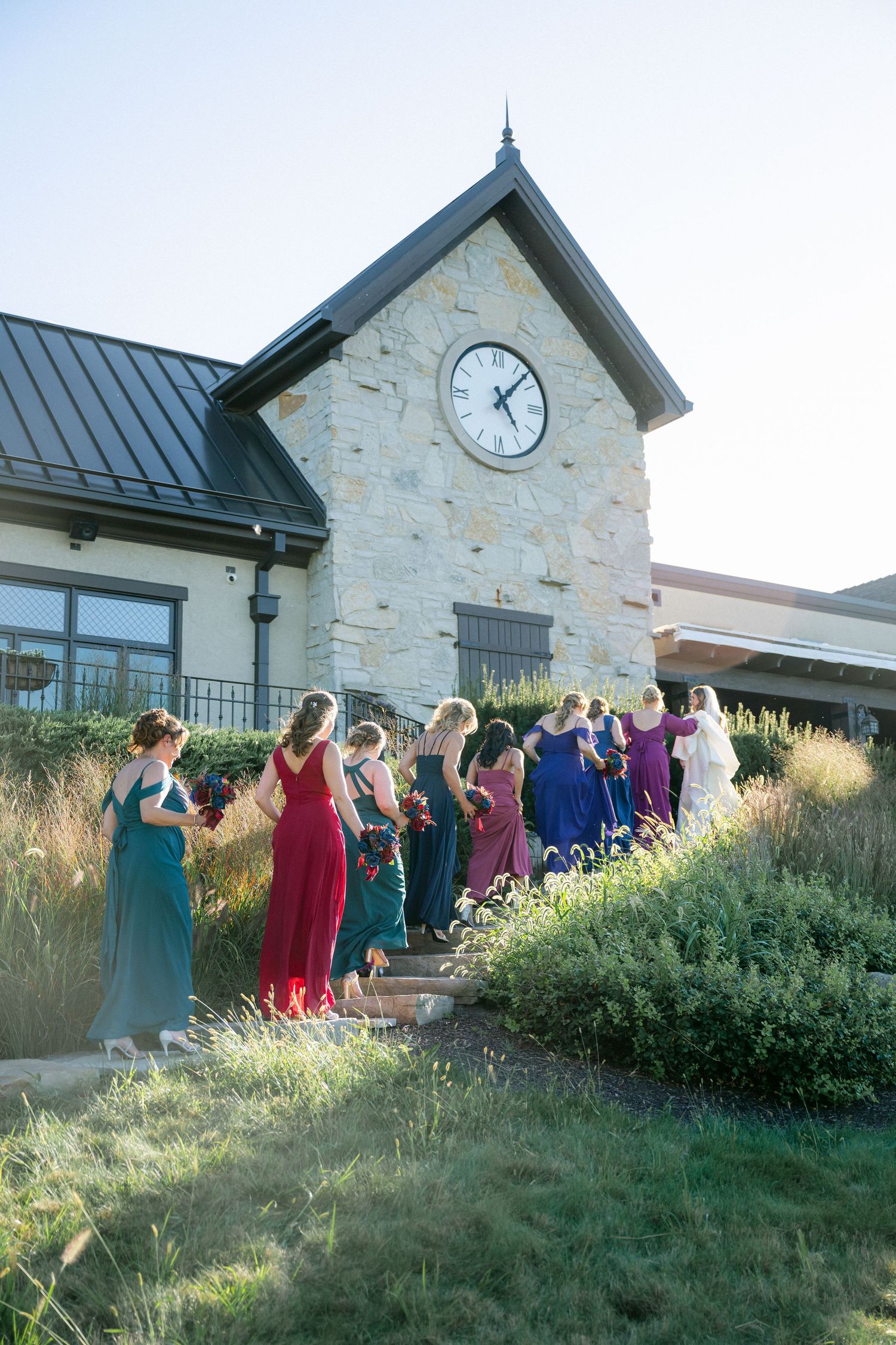 Wedding party in colorful formal attire stands on landscaped hillside by stone clock tower building at sunset.