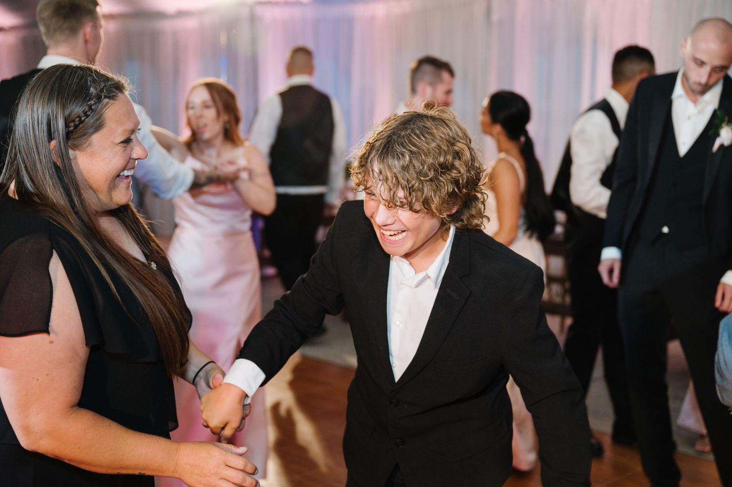 Wedding guests dance and socialize at an indoor reception with white draping in the background.