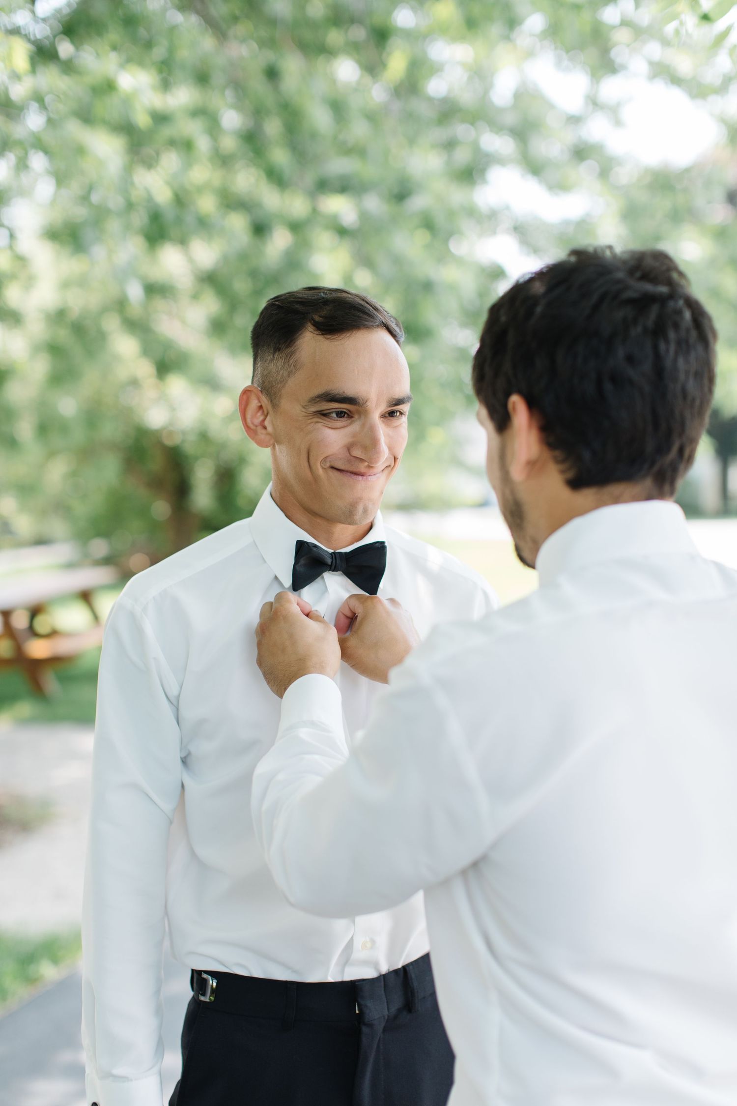 A person helps adjust another's black bow tie against a crisp white dress shirt in an outdoor setting.
