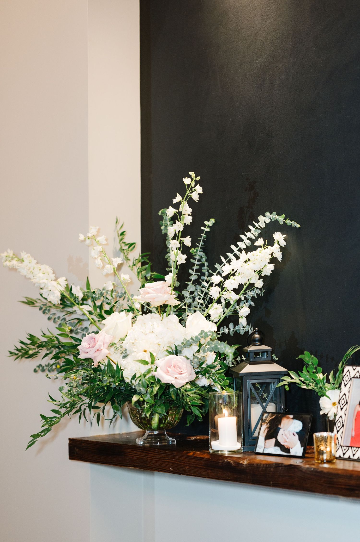 White floral arrangement with roses and greenery sits on a wooden shelf alongside a lantern and decorative items.